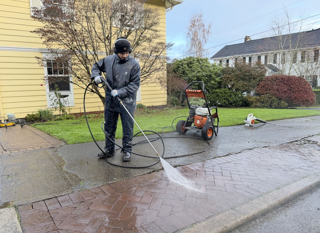Person wearing protective clothing using a pressure washer to clean a brick driveway in front of a yellow house.