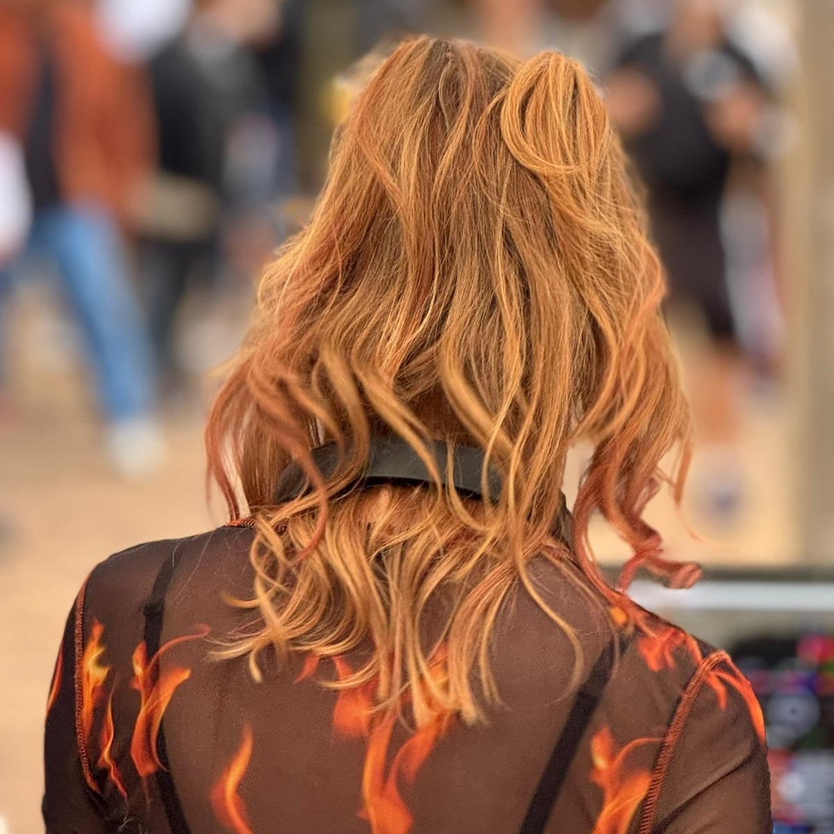 Person mit lockigen, roten Haaren und durchsichtigem schwarzen Oberteil mit Flammenmuster von hinten aufgenommen.