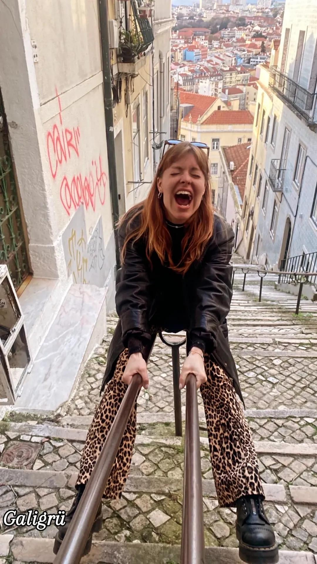Frau mit Leopardenhose und schwarzem Mantel lacht und hält sich an Handläufen einer steilen Treppe in der Stadt fest.