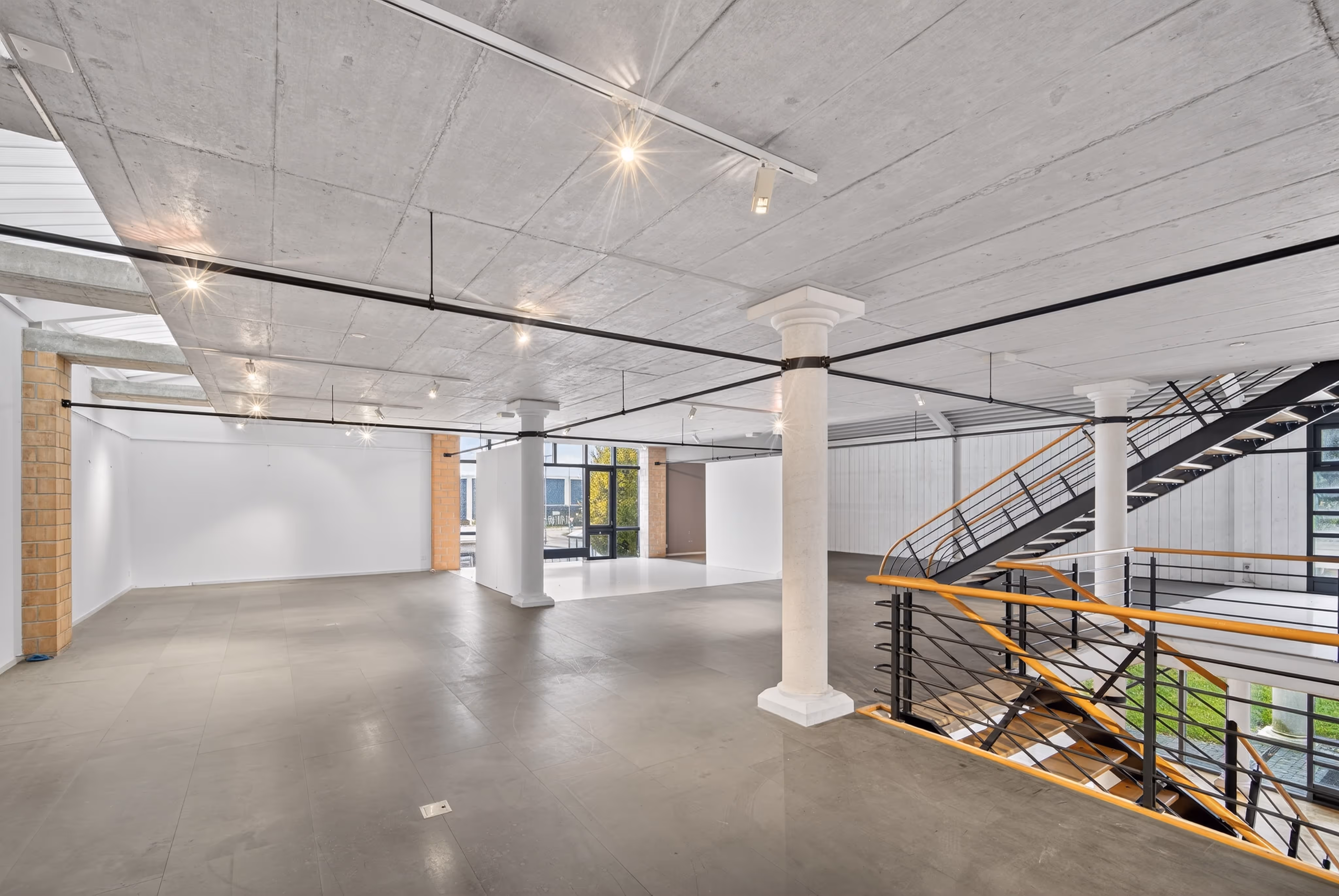Spacious empty modern loft with concrete ceiling, white columns, large windows, and a black metal staircase with wooden handrails.