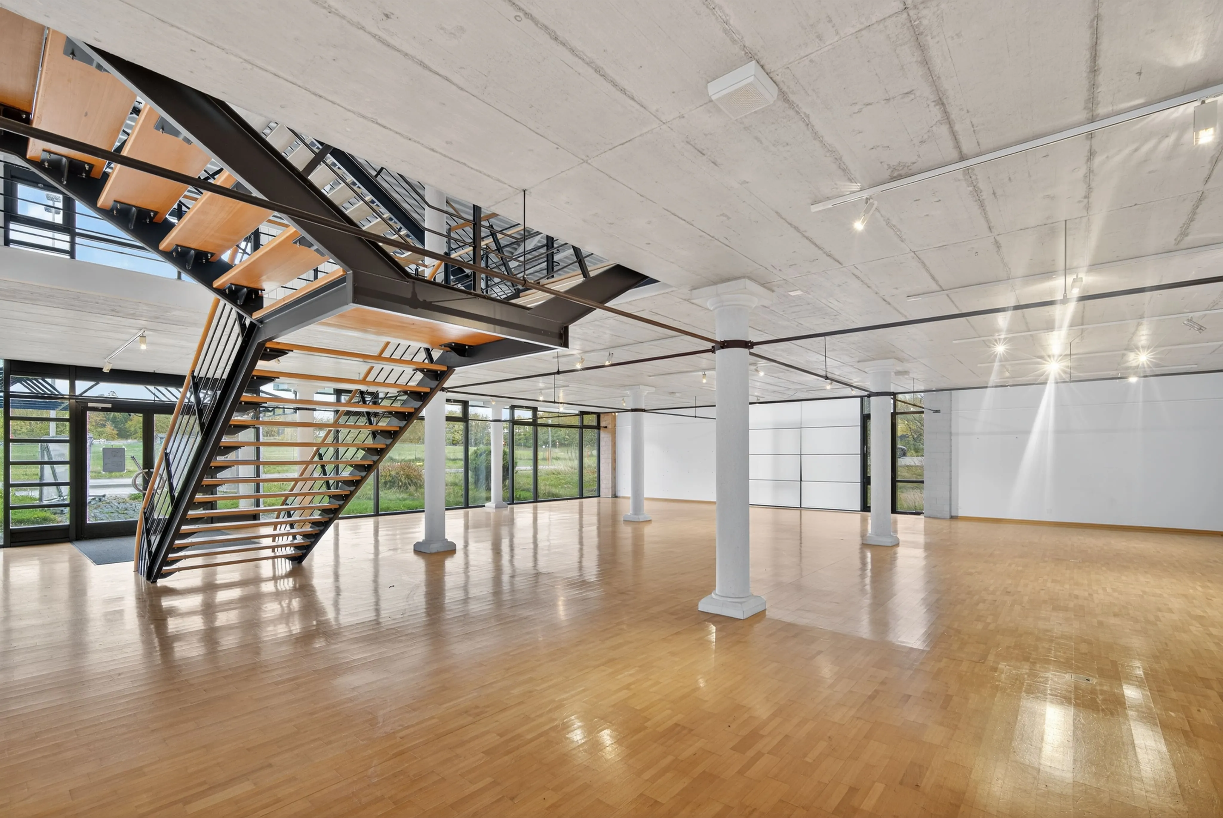 Spacious modern empty room with polished wooden floor, large glass windows, white pillars, and a black metal staircase with wooden steps.
