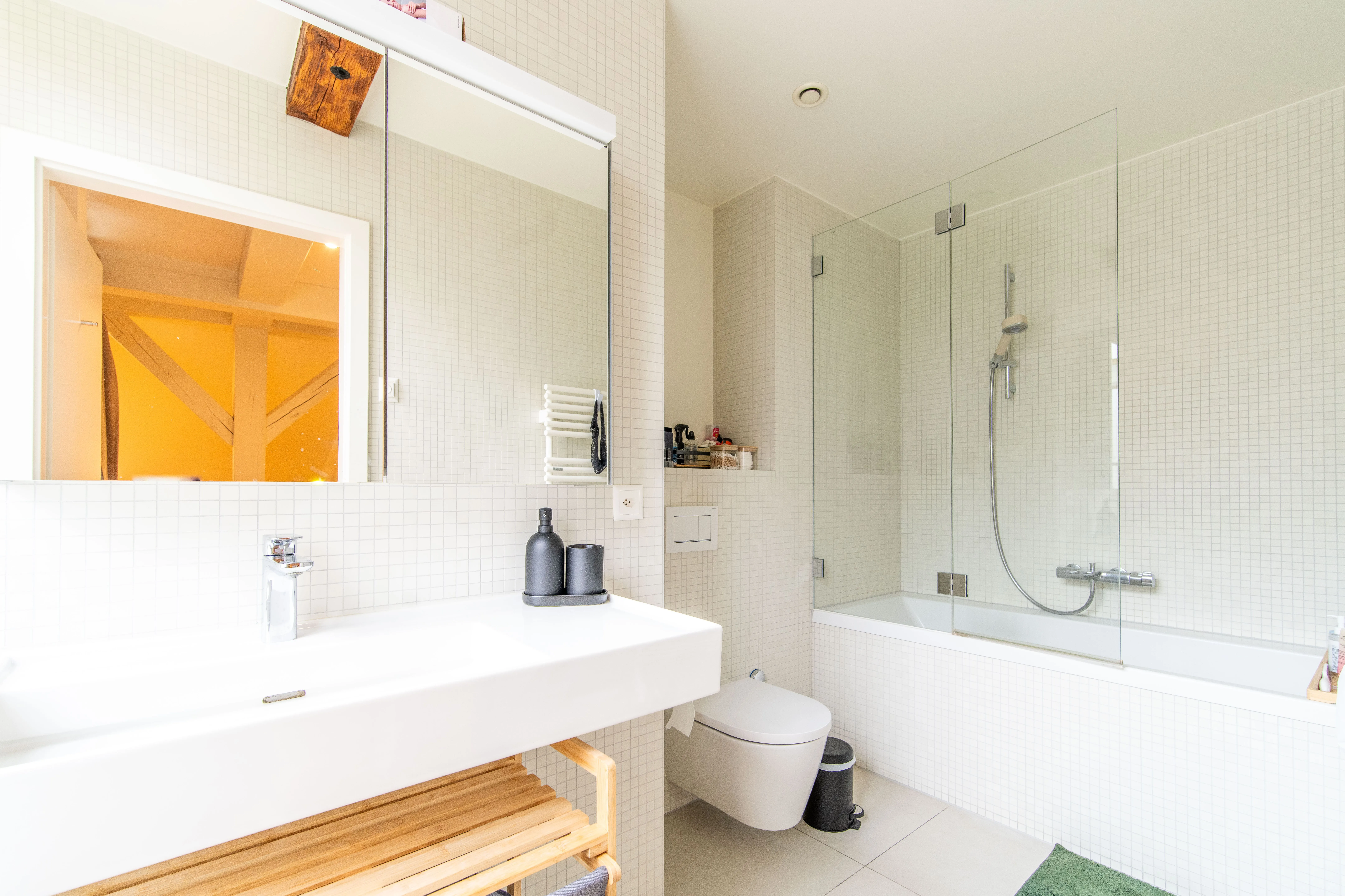Modern bathroom with white tiled walls, a wall-mounted toilet, bathtub with glass shower screen, large sink, and wooden shelf underneath.