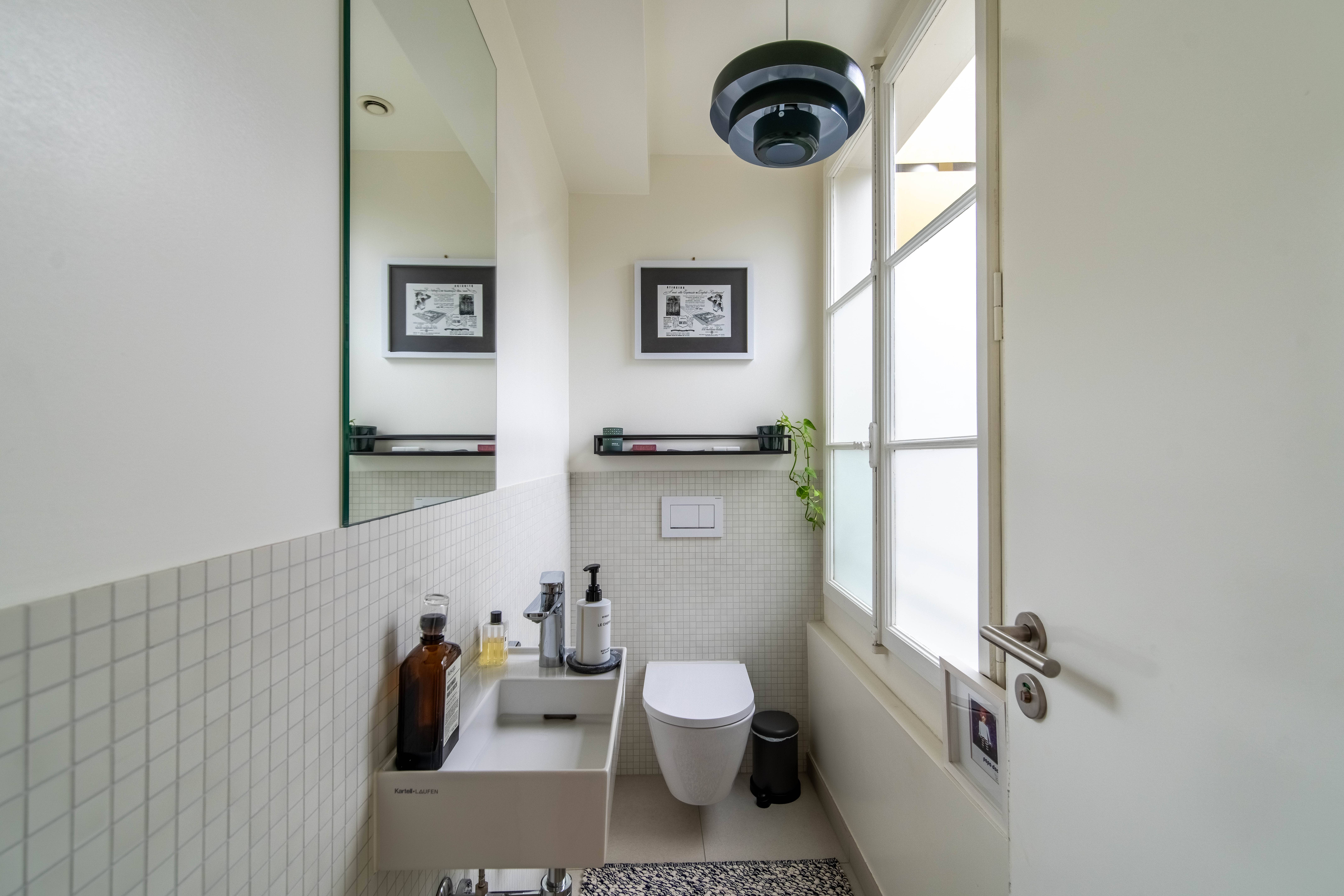 Minimalist bathroom with wall-mounted toilet, small white sink with toiletries, a large mirror, a black trash can, and a framed picture above the toilet.