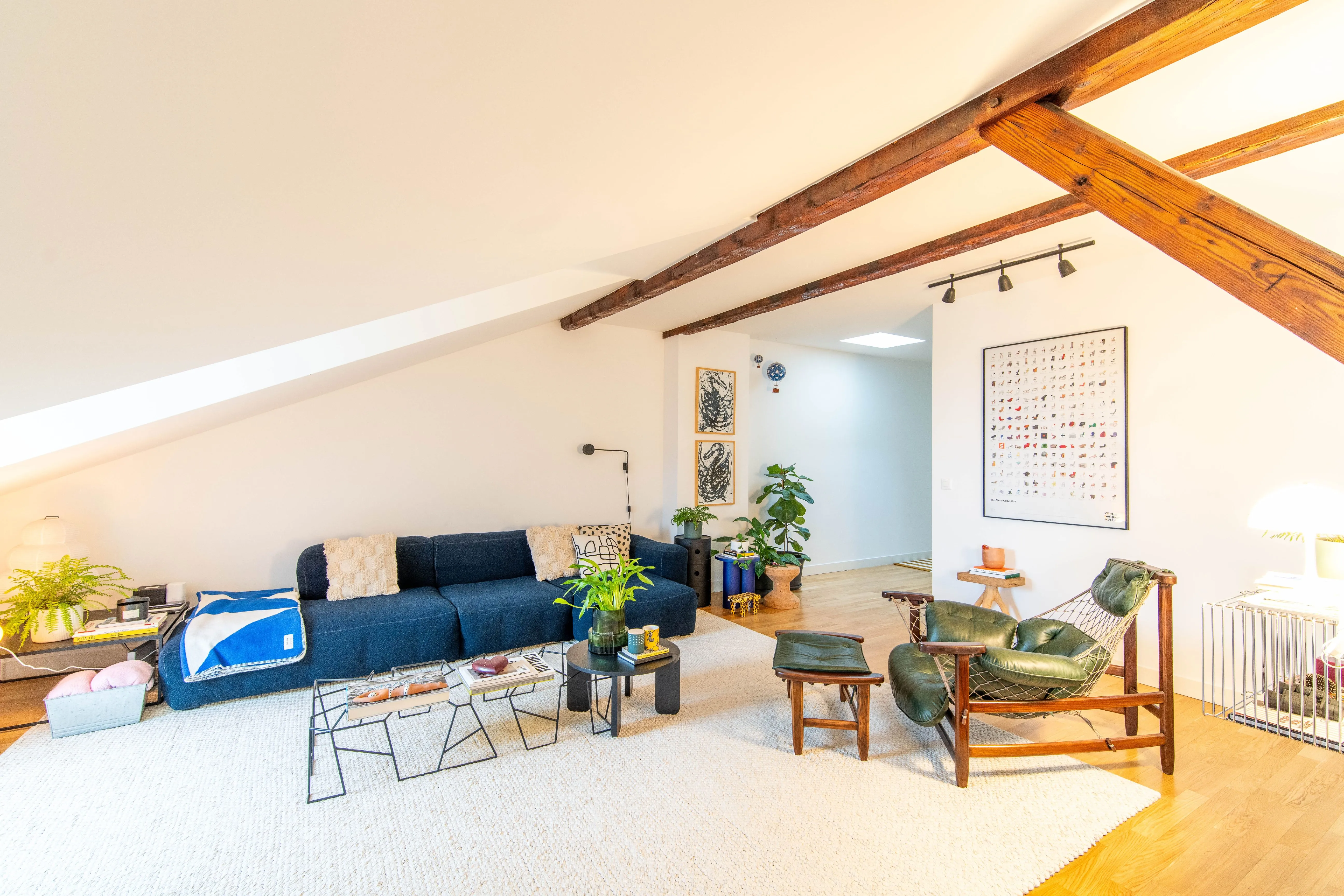 Bright living room with slanted ceiling, dark blue sofa with pillows, green leather armchair with matching footstool, wooden beams, and indoor plants.
