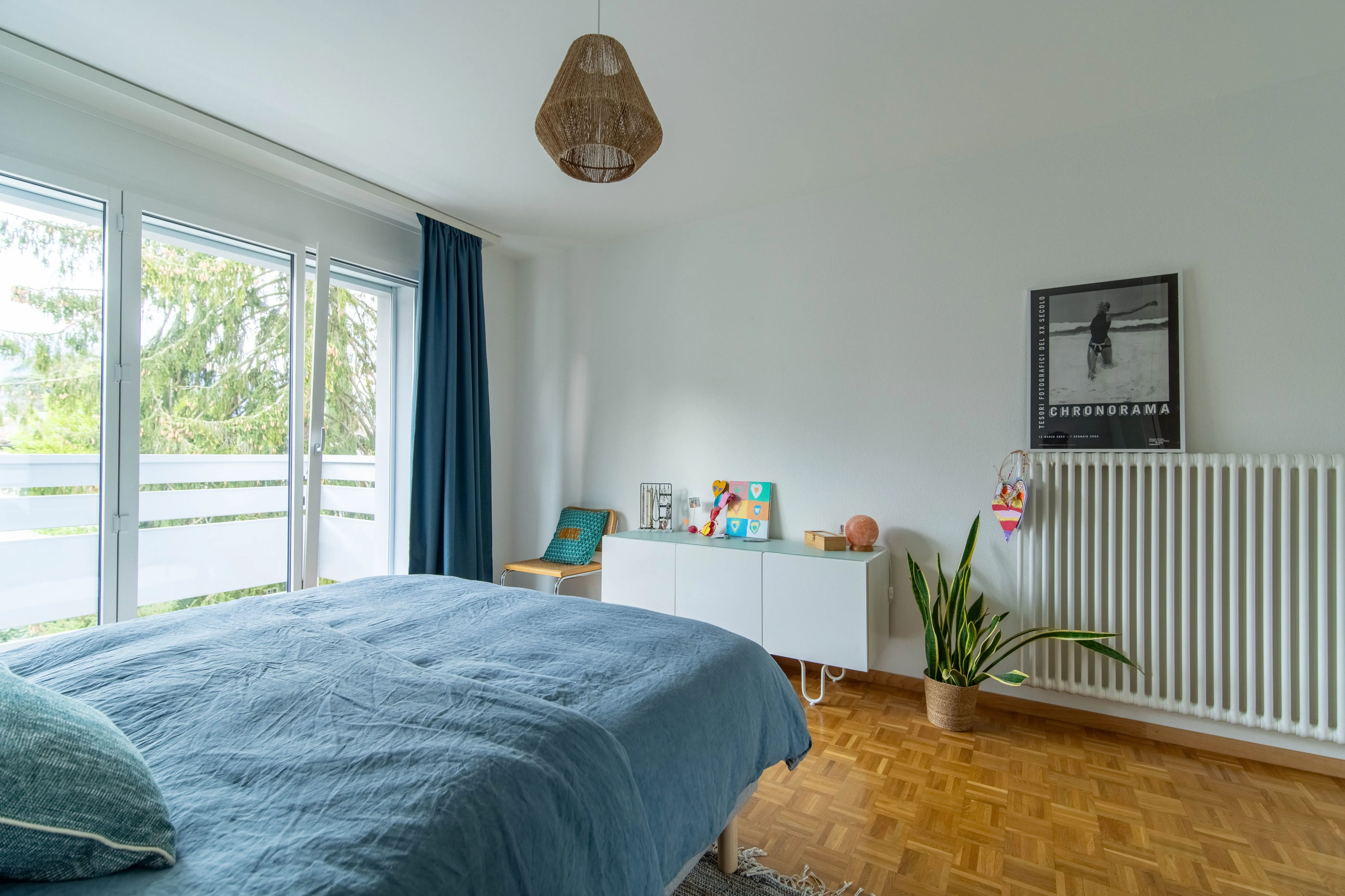 Bright bedroom with large windows, blue bed cover, white cabinet, wooden chair, potted plant, and wall radiator with framed photo above.