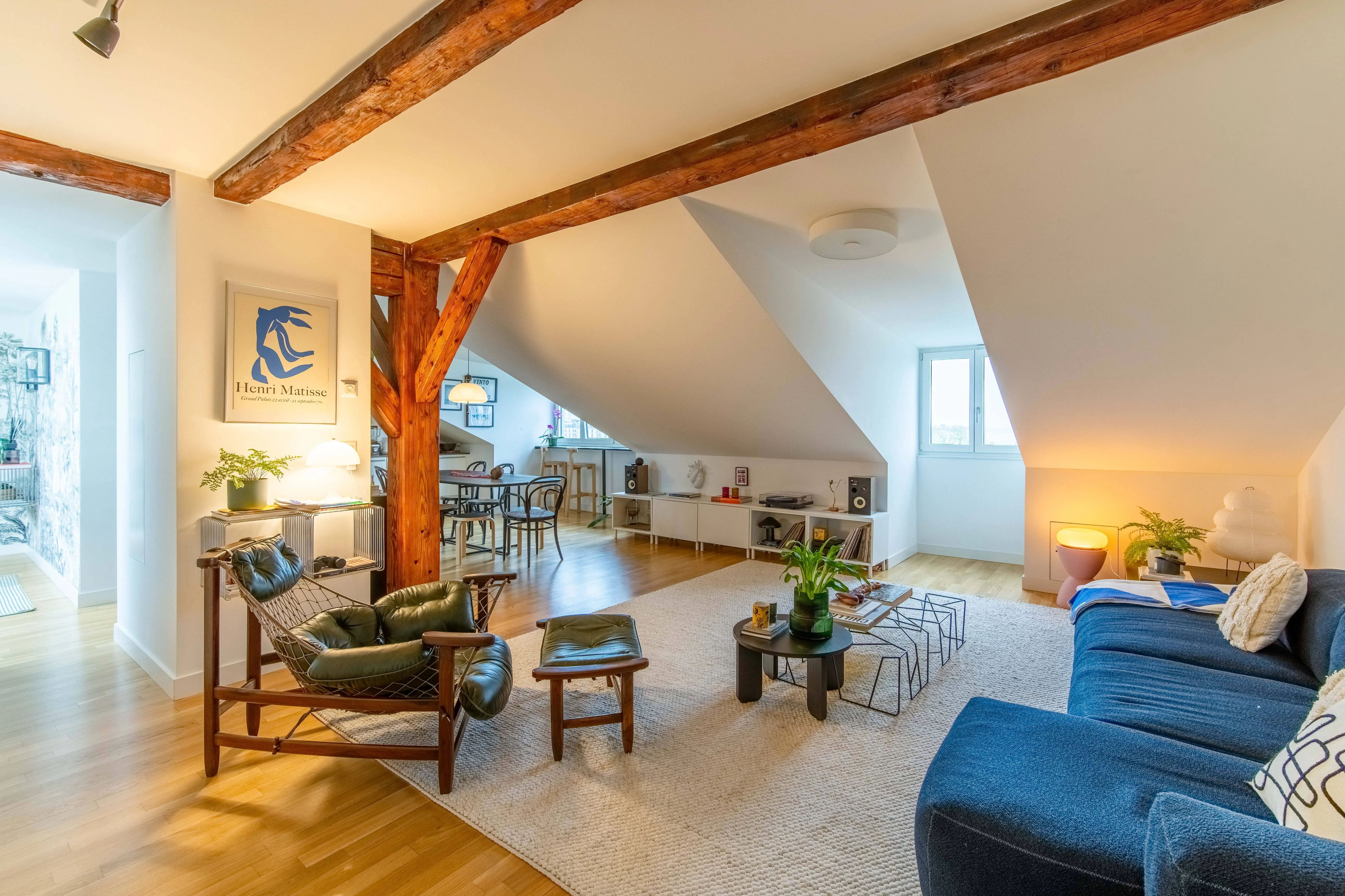 Bright attic living room with slanted ceilings, wooden beams, a green leather chair with matching footstool, a blue sofa, and a round coffee table with plants.