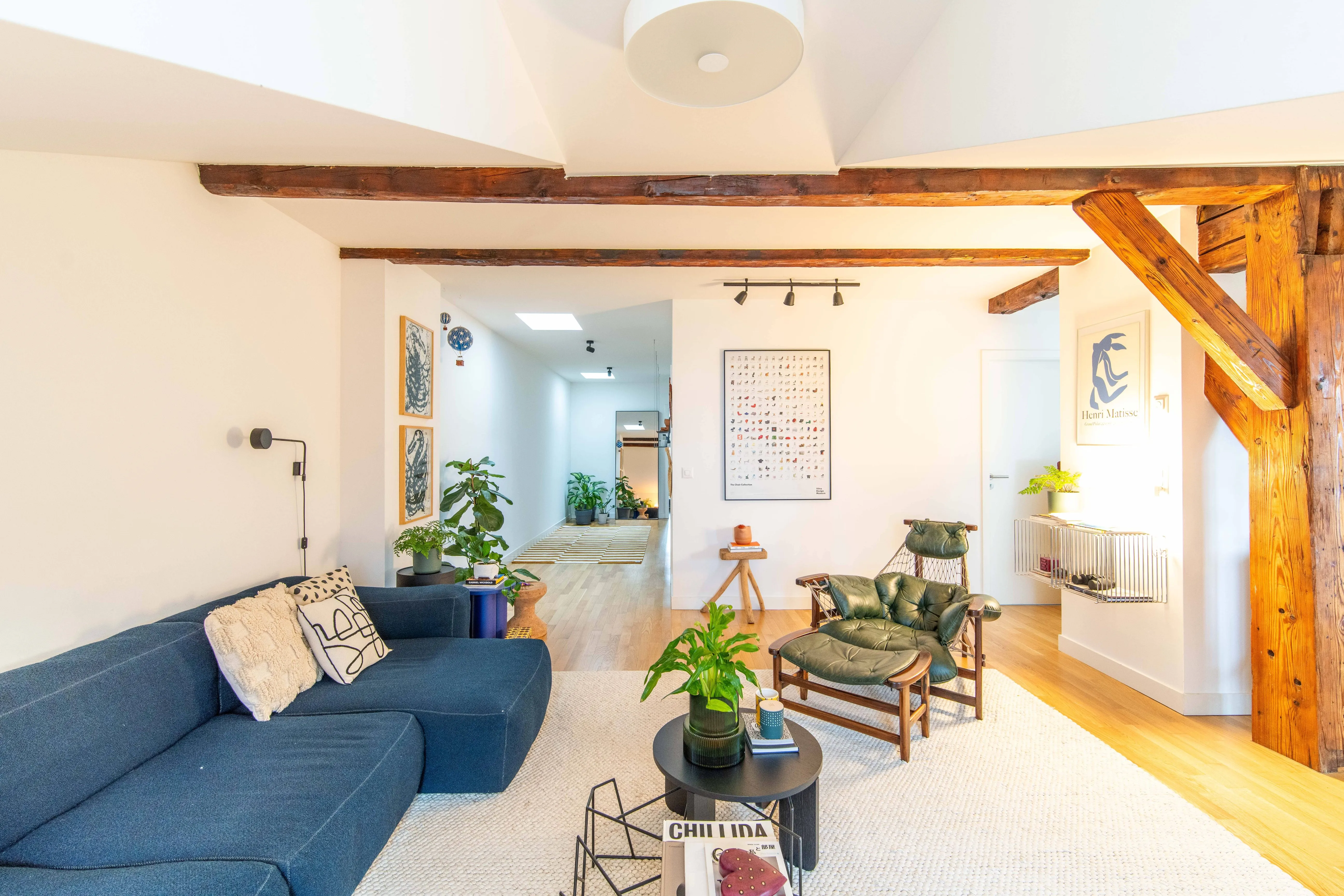 Bright living room with exposed wooden beams, blue sectional sofa, green leather lounge chair, coffee tables with plants, and framed art on white walls.