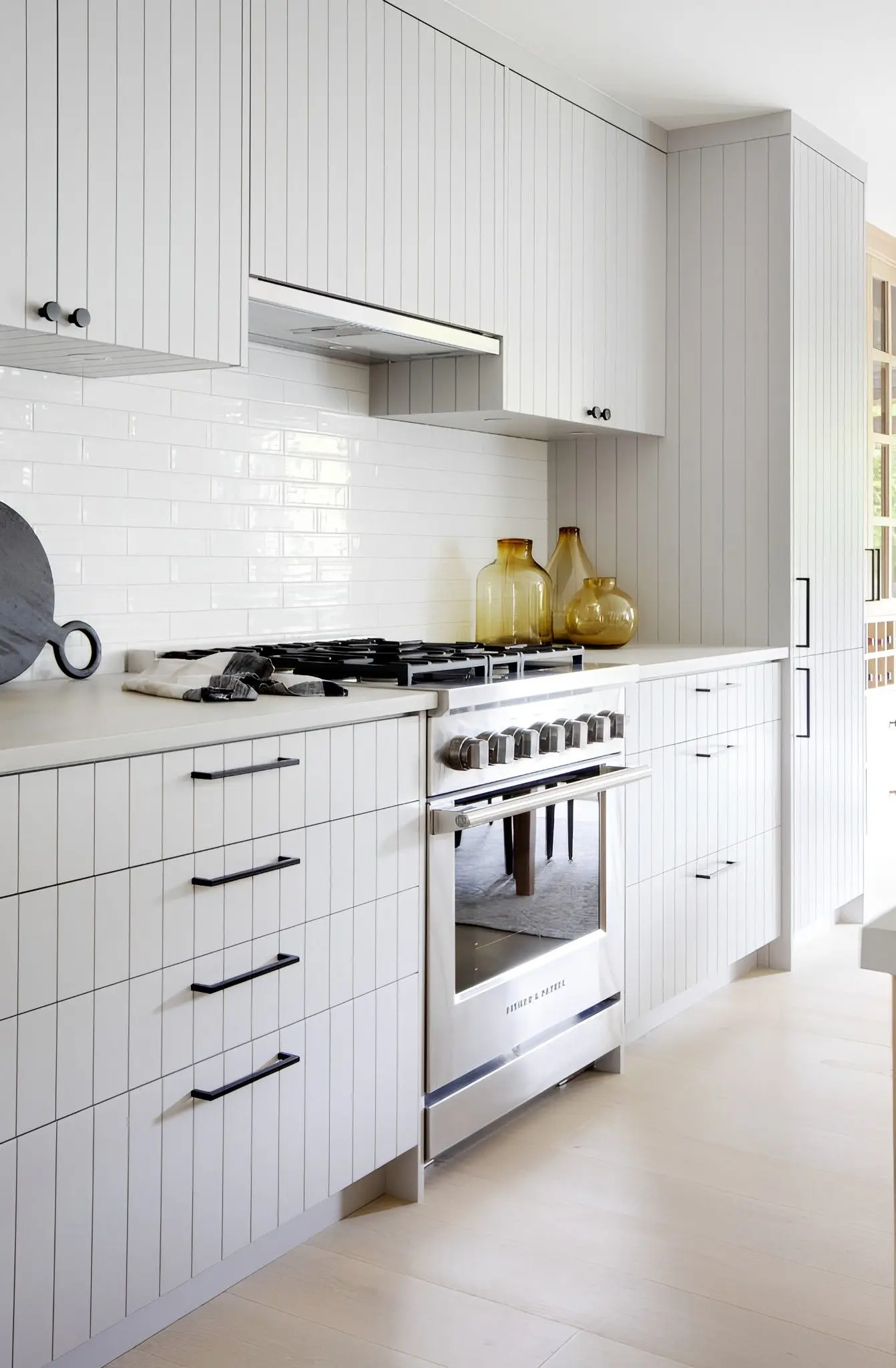 Modern kitchen with white cabinetry featuring vertical lines, black handles, a stainless steel gas stove, and amber glass decorative bottles on the counter.