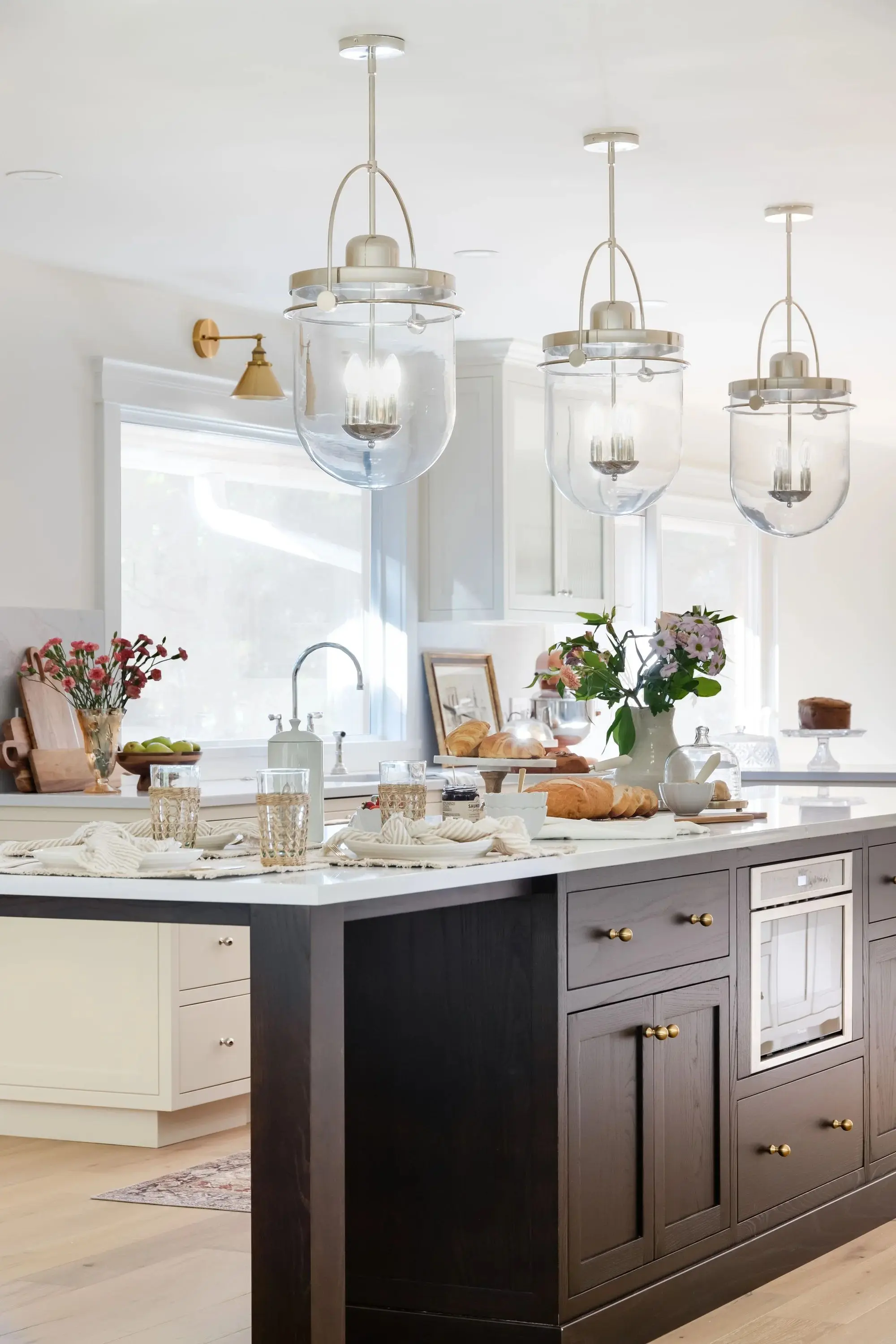 Modern kitchen with dark wood island, white marble countertop, pendant lights, flowers, bread, and glassware.