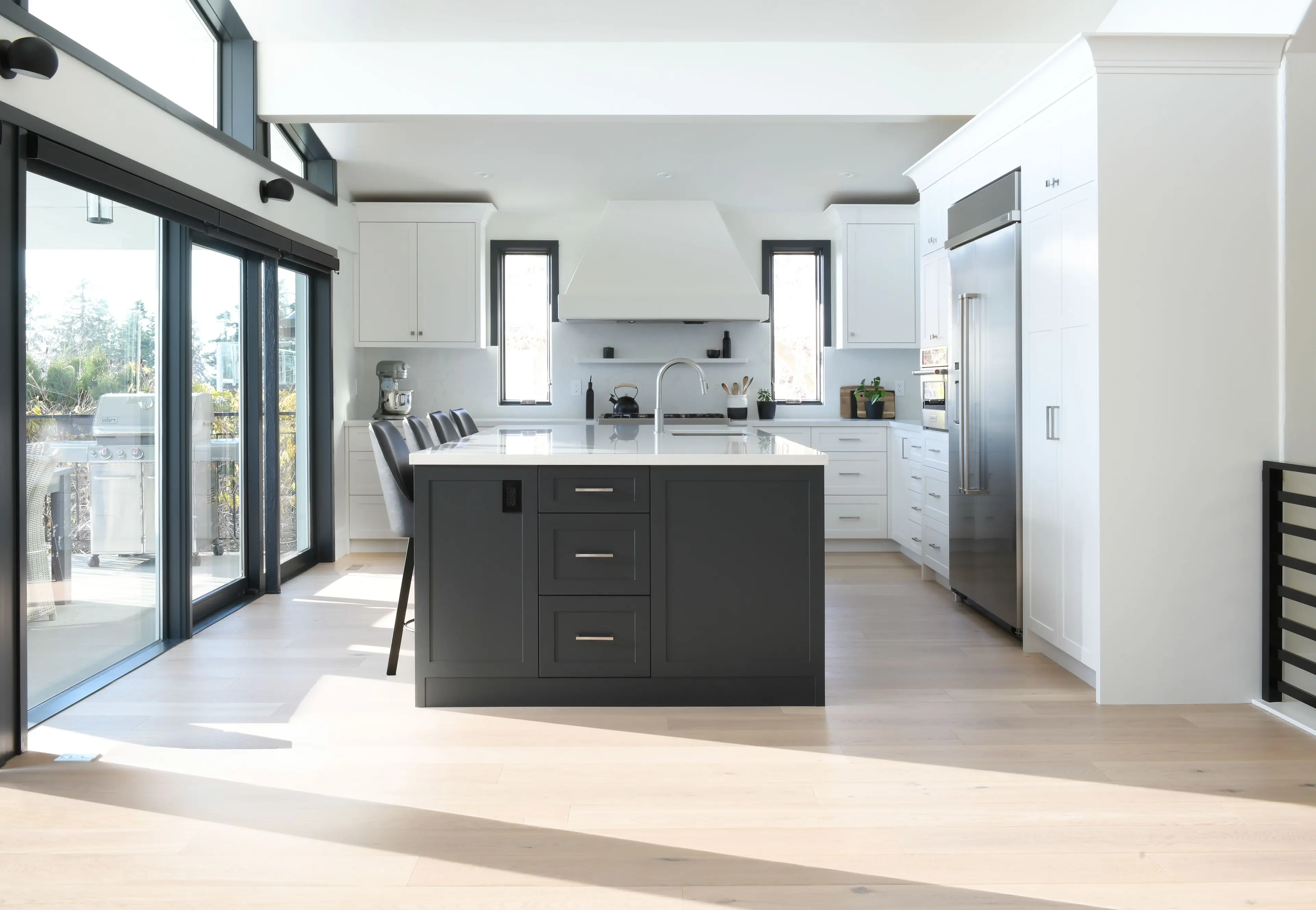 Bright modern kitchen with a large dark gray island, white cabinets, stainless steel refrigerator, and sliding glass doors leading outside.