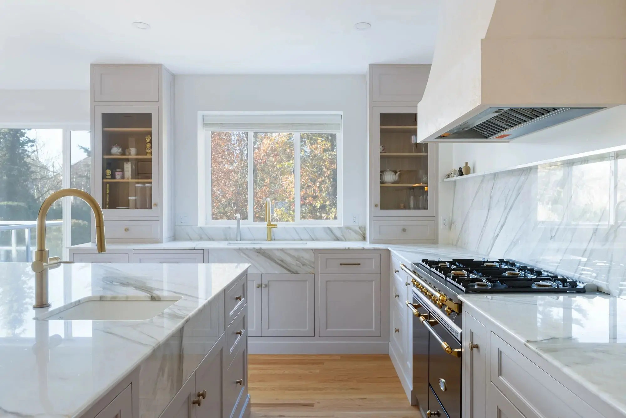 Bright modern kitchen with marble countertops, gold fixtures, and a large window over the sink.