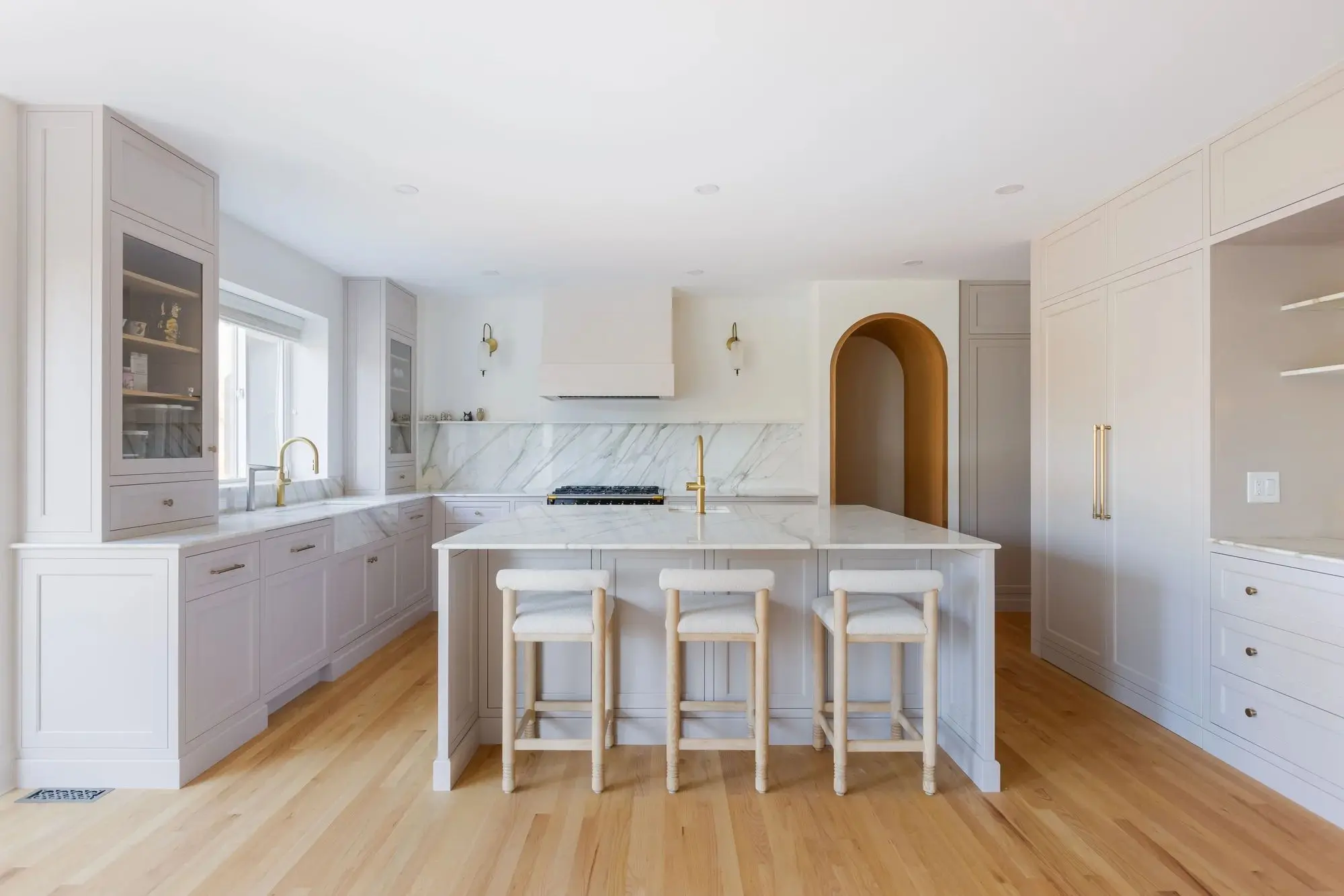 Modern kitchen with white cabinetry, marble countertops and backsplash, three padded stools at central island, and wooden flooring.