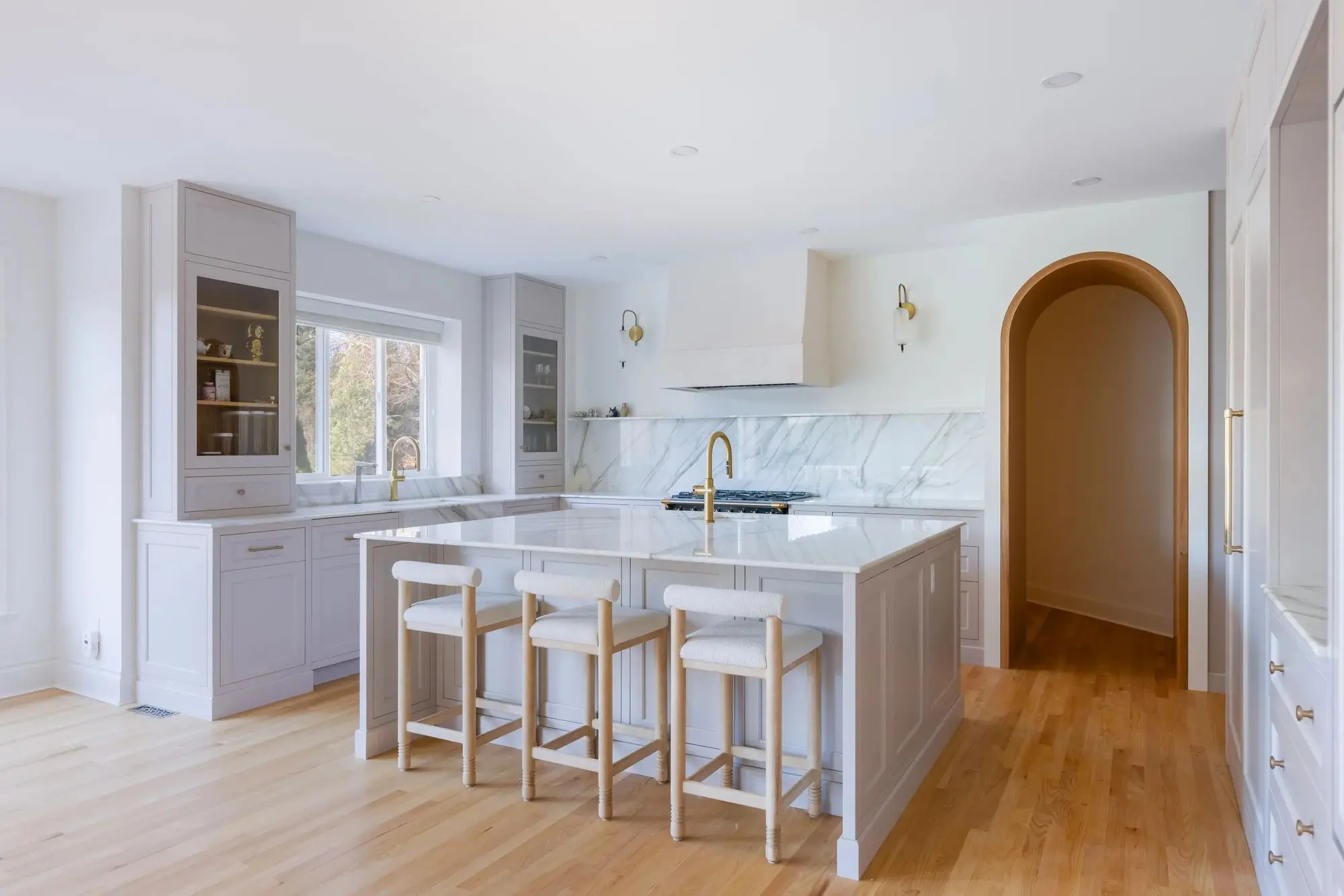 Bright modern kitchen with light wood flooring, white cabinetry, marble countertops, a kitchen island with three upholstered stools, and gold fixtures.