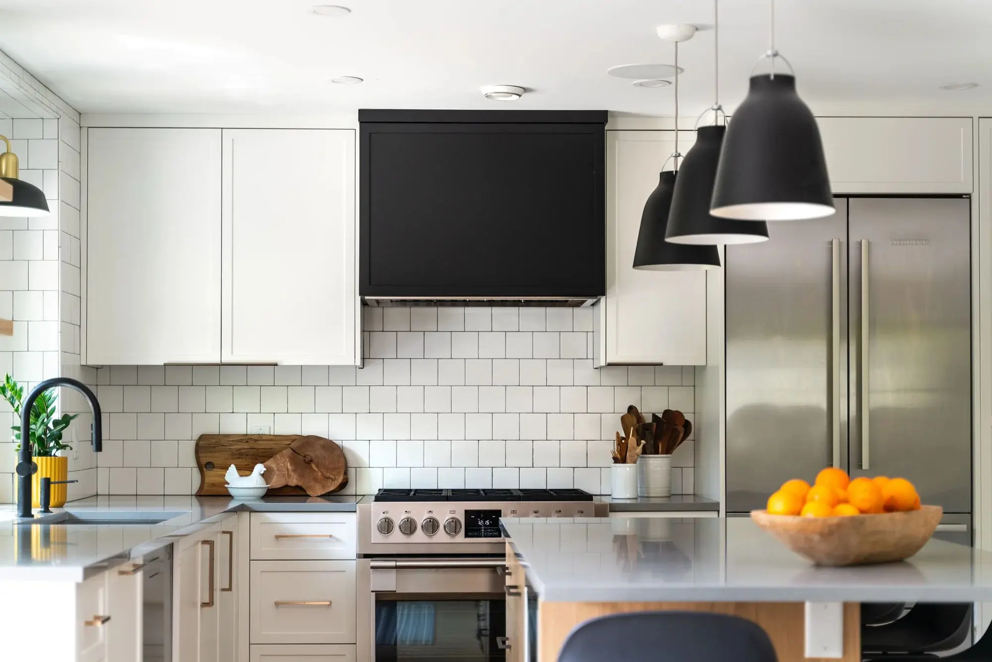 Modern kitchen with white cabinetry, black range hood, stainless steel stove and refrigerator, black pendant lights, and a bowl of oranges on the island.
