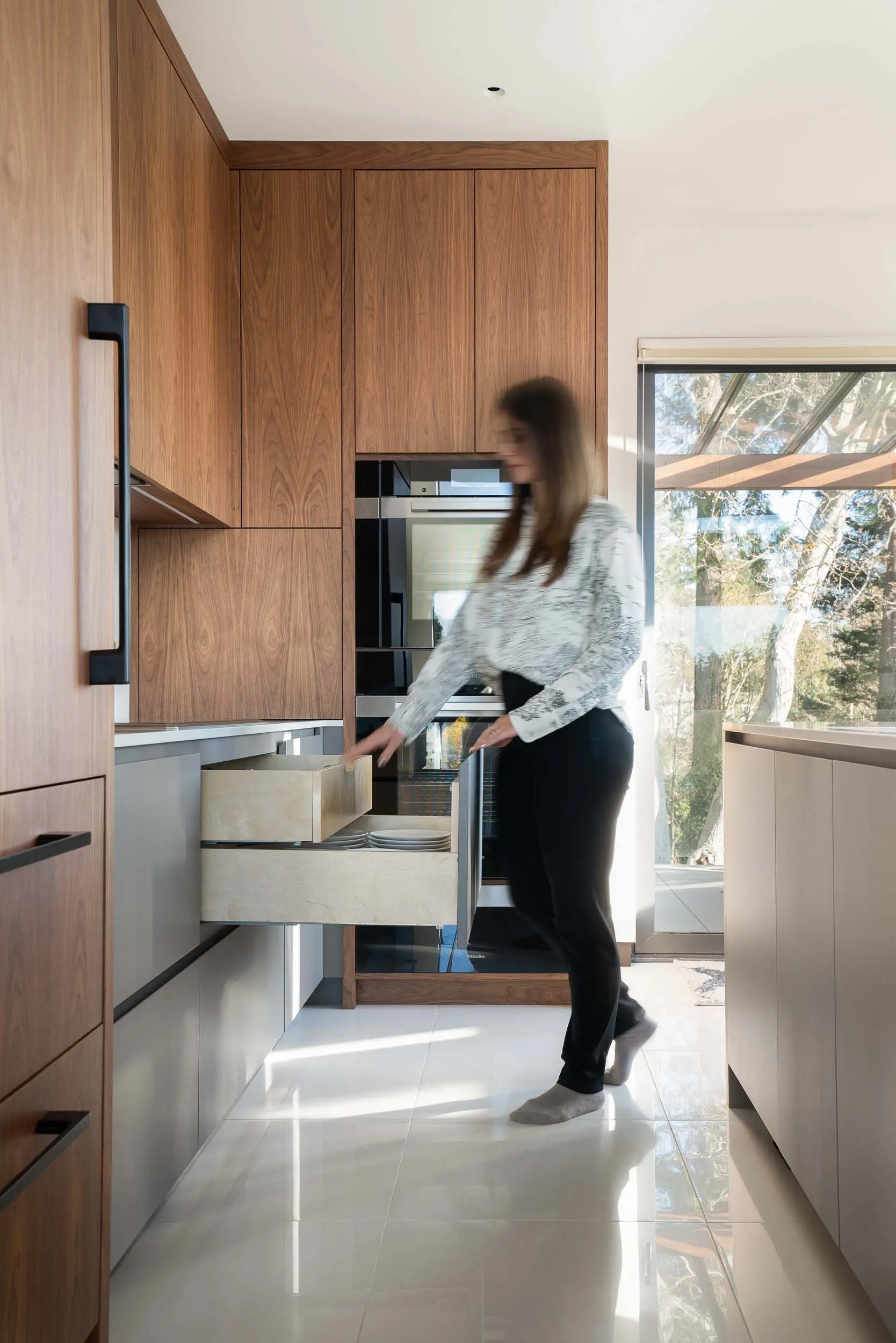 Woman opening wooden kitchen drawers in a modern kitchen with natural light coming through a glass door.