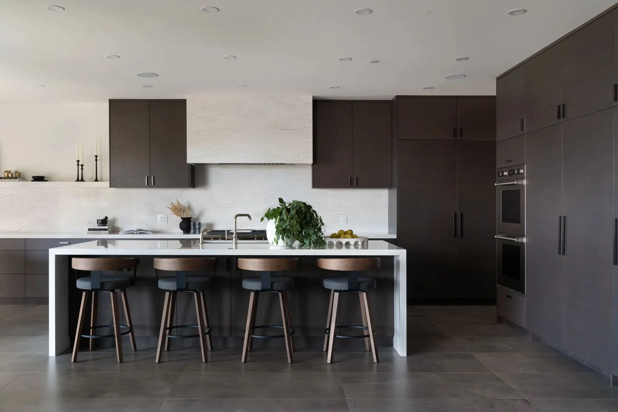 Modern kitchen with dark cabinetry, a white island with four wooden stools, a sink, potted plant, and fruit bowl.