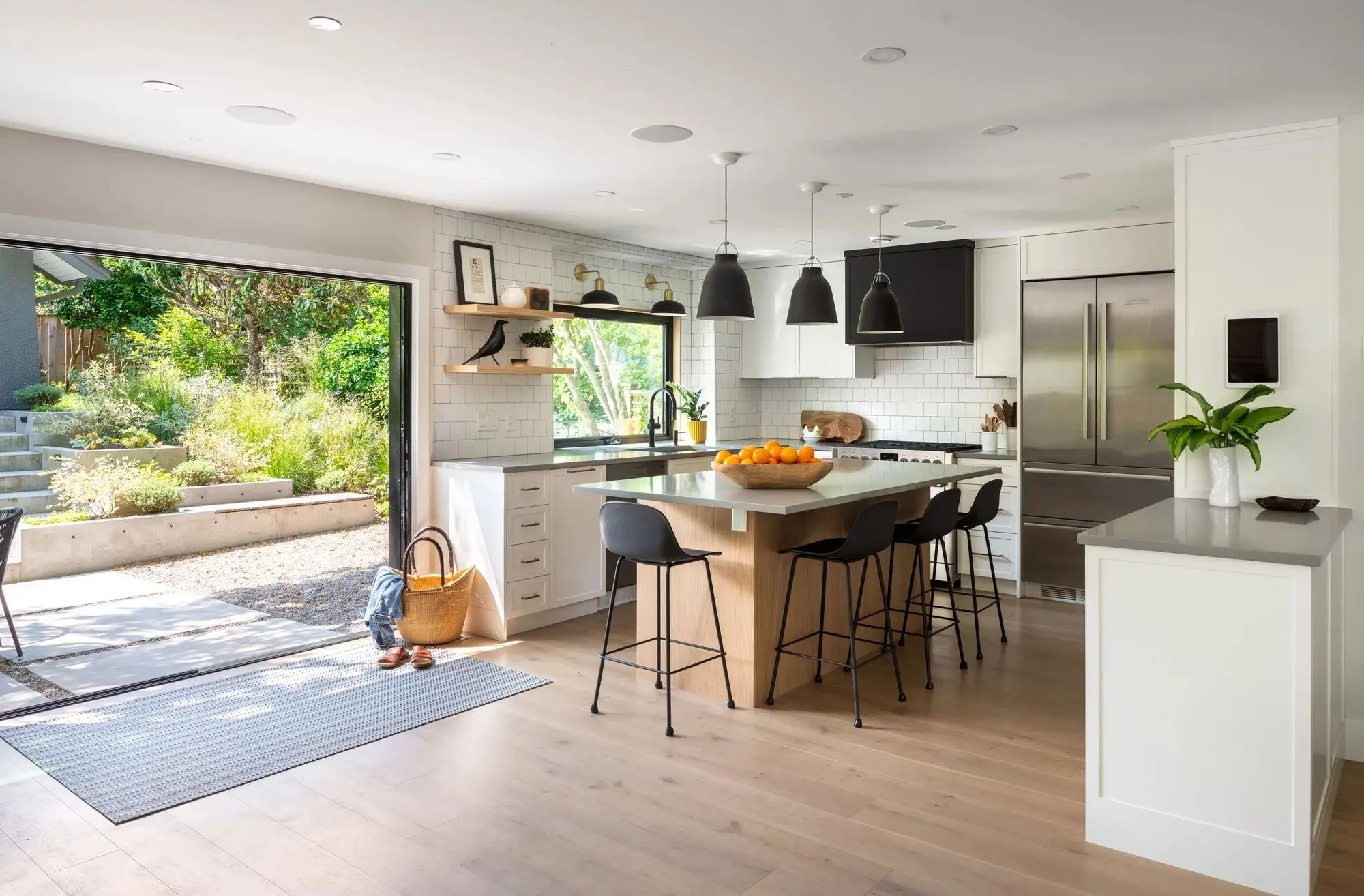 Bright modern kitchen with a large island, black bar stools, black pendant lights, stainless steel refrigerator, white cabinets, and open doors leading to a green backyard.