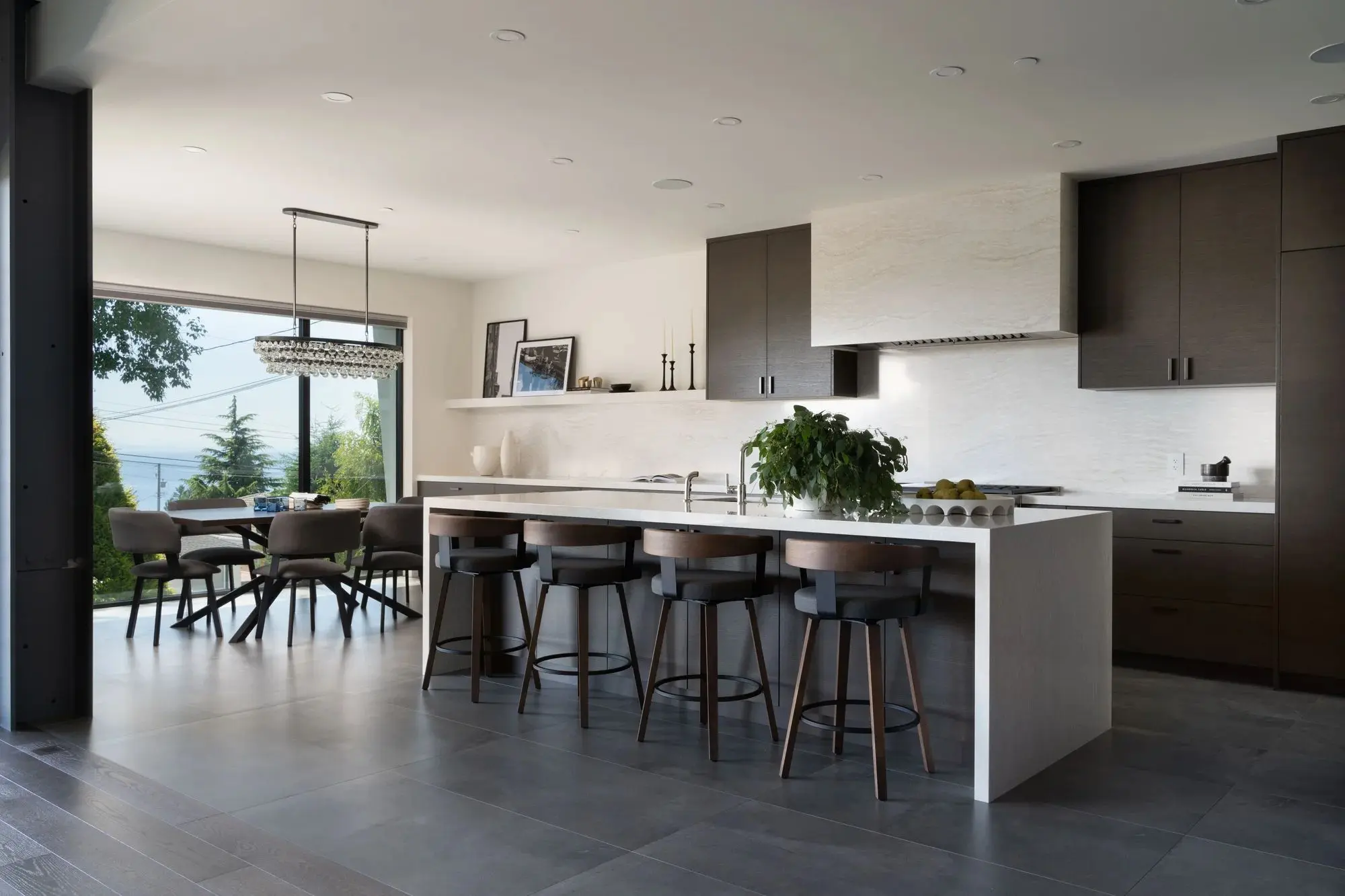 Modern kitchen with a white island and four dark wood bar stools, next to a dining area with a round table and chairs near large windows overlooking greenery.