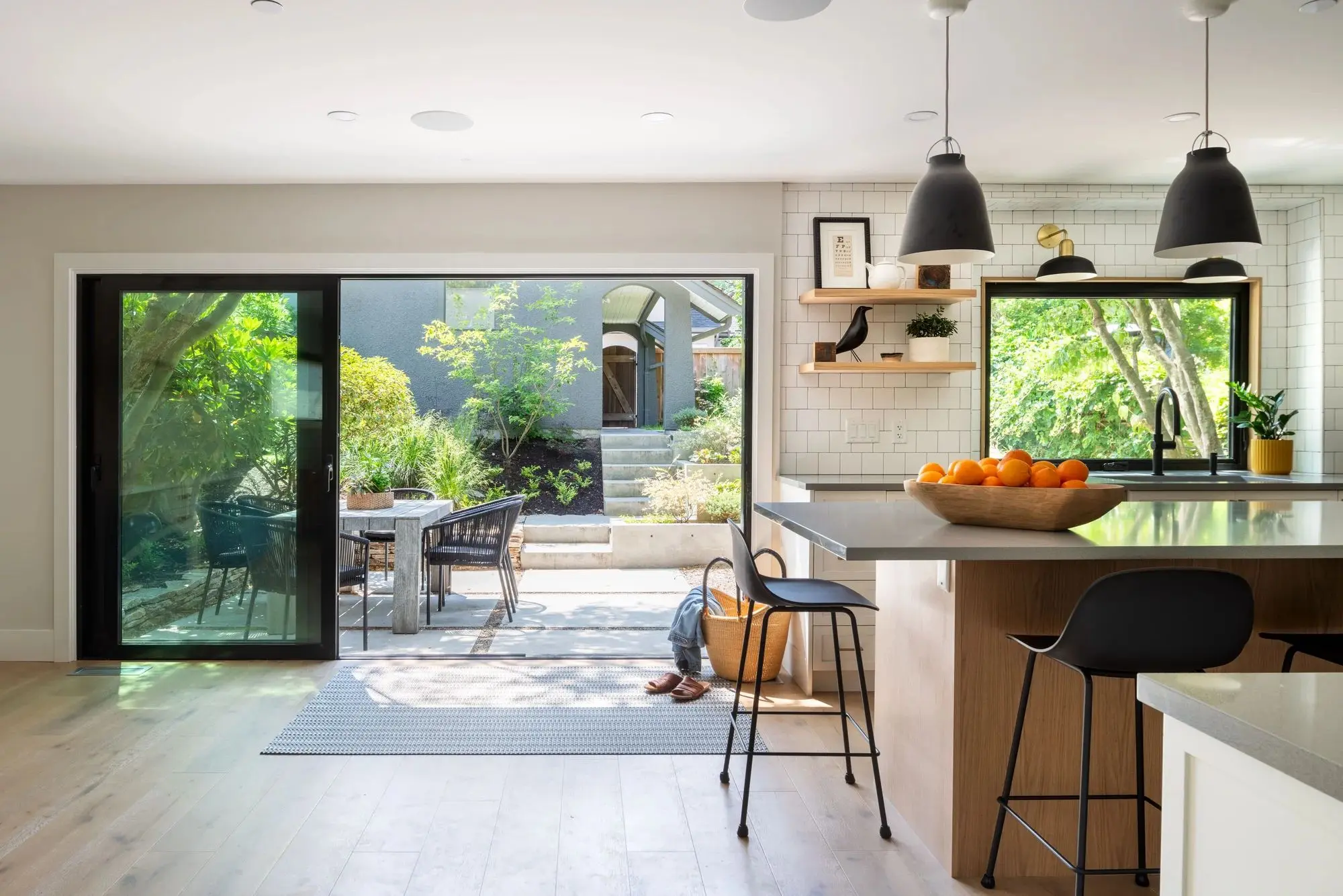 Bright modern kitchen with large island, black bar stools, hanging pendant lights, and sliding glass door opening to a green backyard patio.