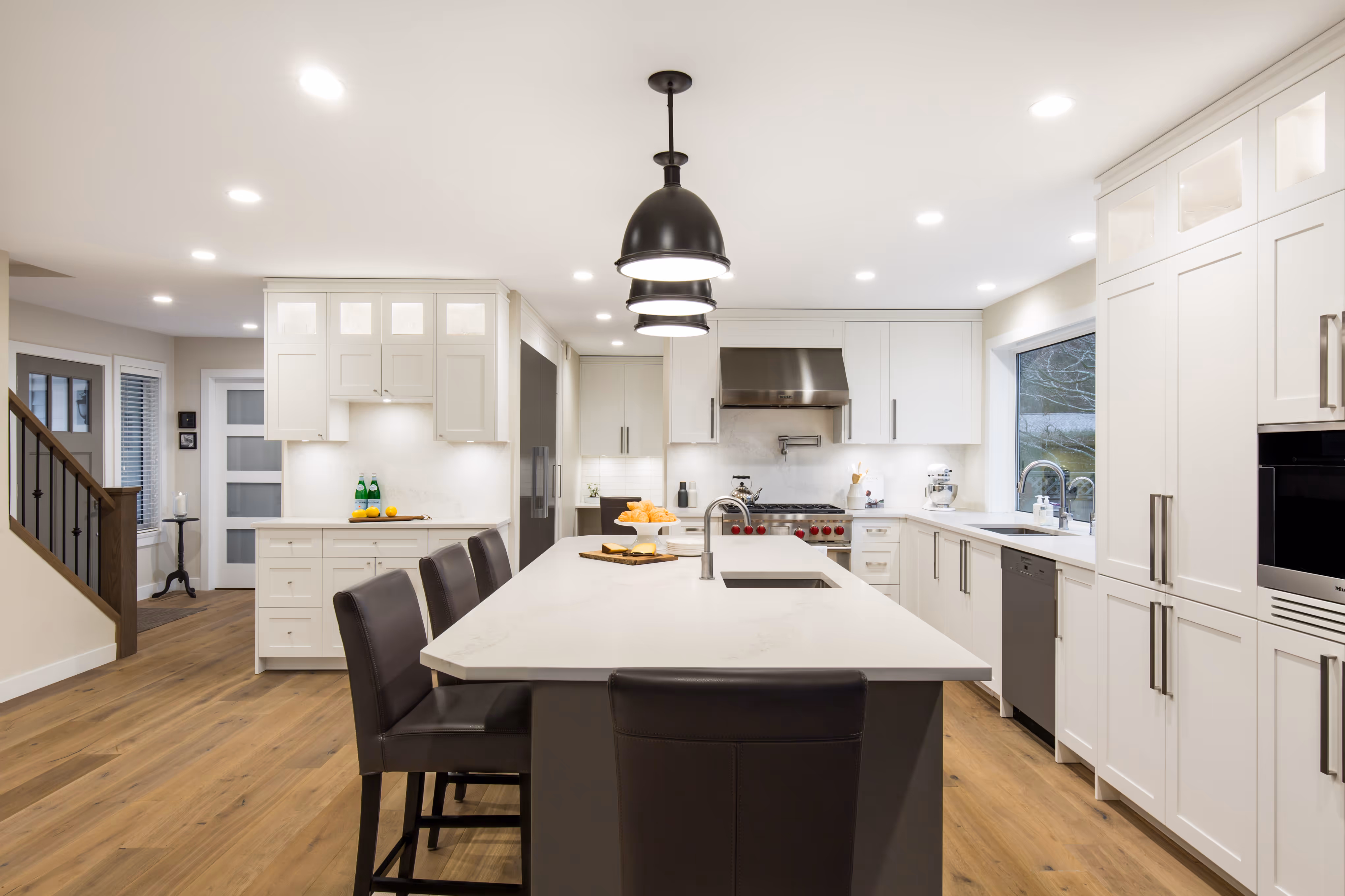 Modern kitchen with white cabinetry, a large island with black leather stools, stainless steel appliances, and wood flooring.