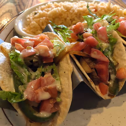 Three tacos filled with beef, lettuce, tomatoes, and drizzled with sauce, served with a side of Mexican rice on a plate. The dish looks appetizing and colorful.
