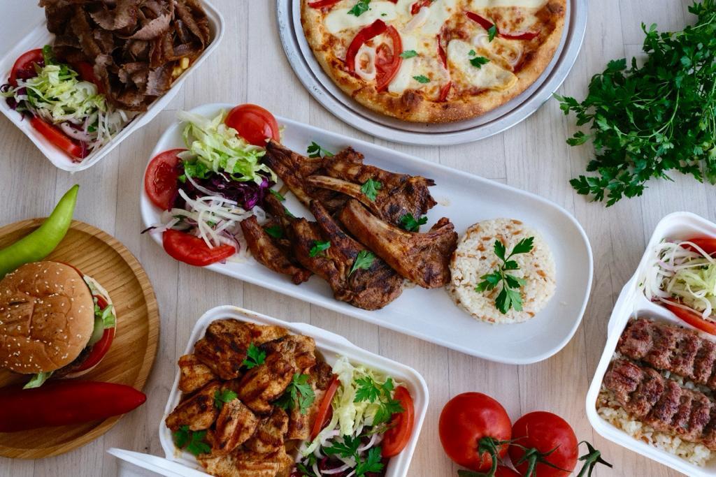 Assorted grilled dishes including lamb chops with rice, grilled chicken, kebabs, a burger, salad, pizza, and fresh tomatoes on a light wooden table.