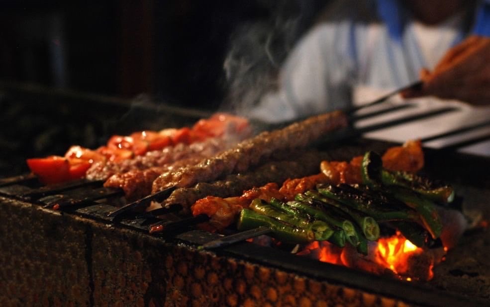 Skewers of grilled meat, green vegetables, and tomatoes cooking over glowing charcoal on a grill.