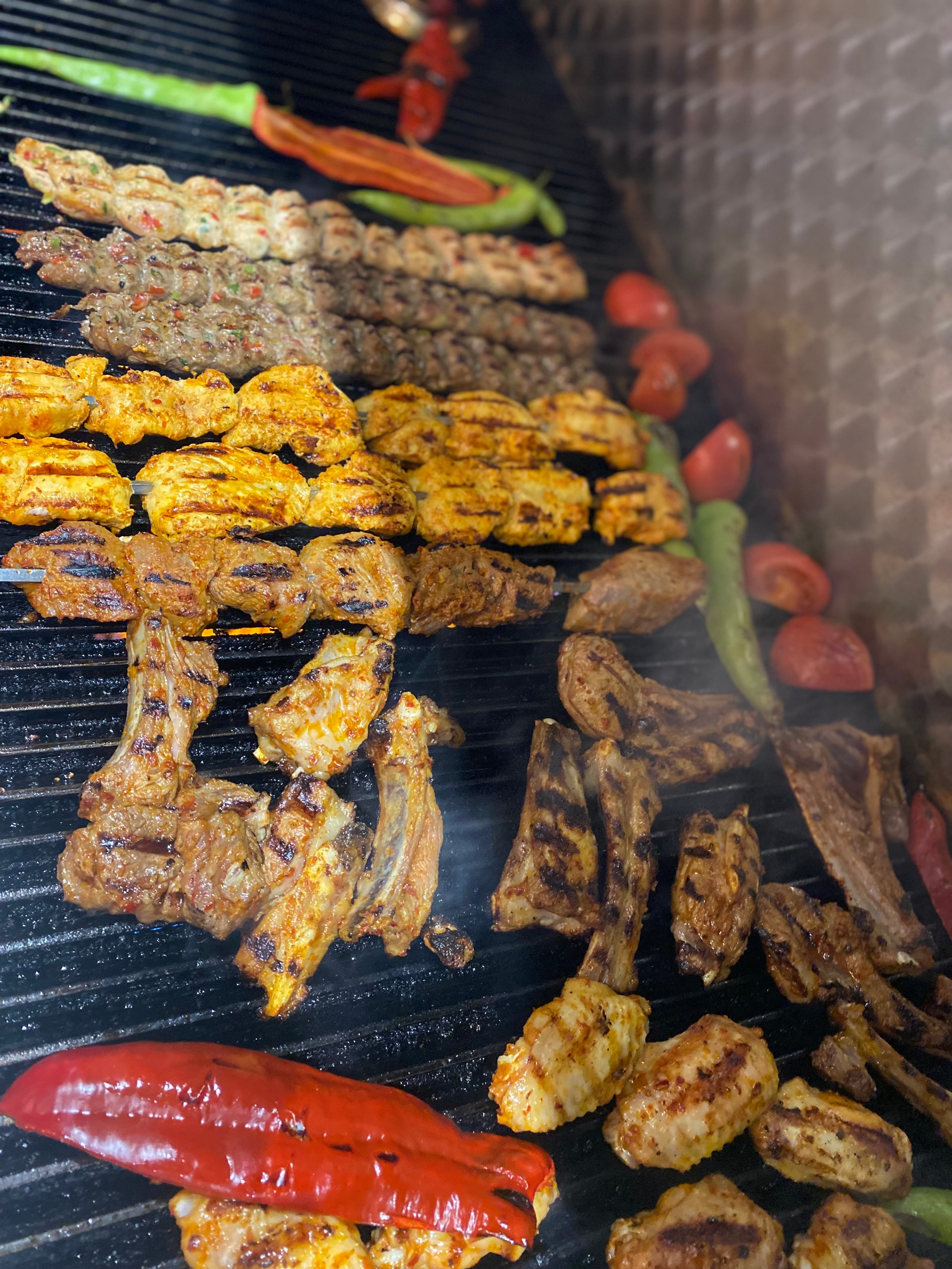 Grill loaded with various grilled meats and vegetables including skewers, ribs, and whole peppers.