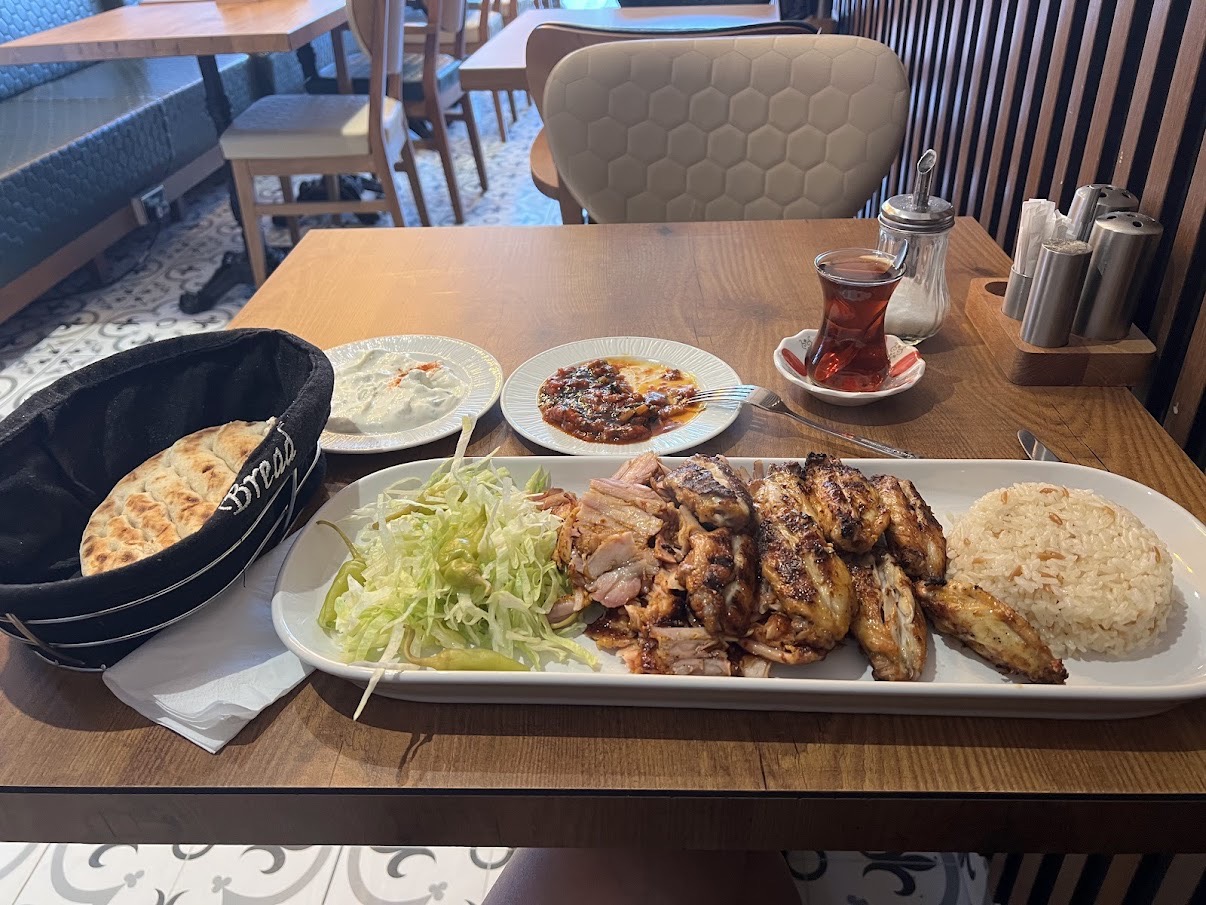 Grilled chicken wings and sliced meat with shredded lettuce and pepper, served with rice, flatbread, two sauces, and a glass of tea on a wooden table in a restaurant.