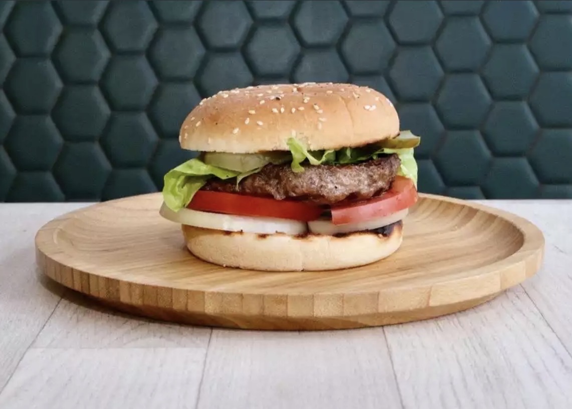 Hamburger with lettuce, tomato, pickles, onion, and a sesame seed bun on a wooden plate.