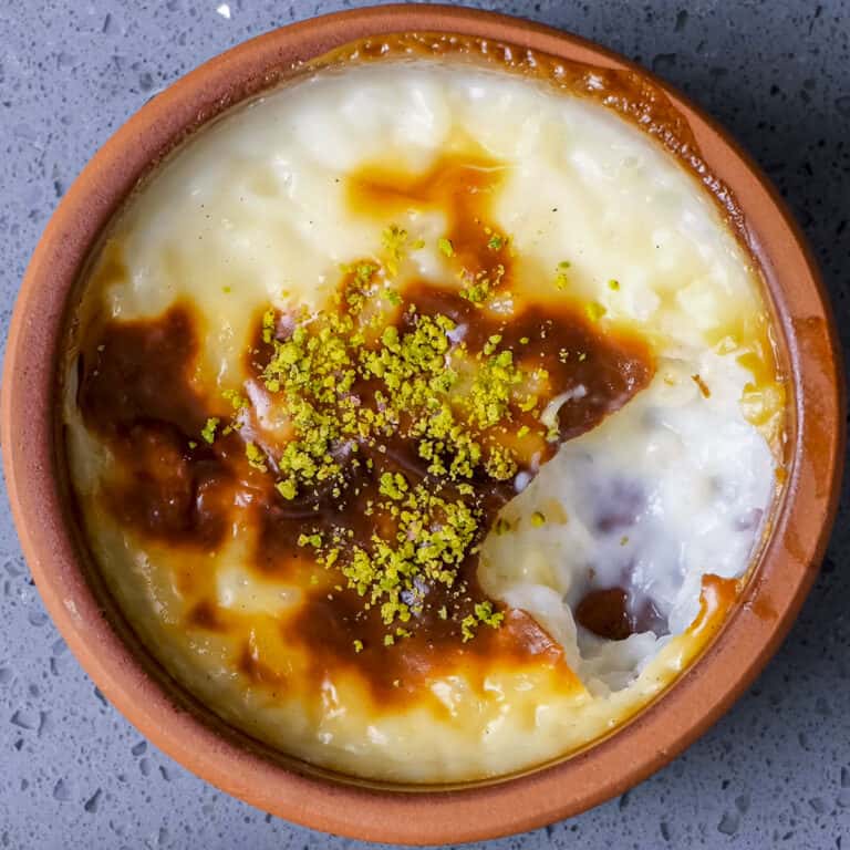 Top view of a baked rice pudding dessert in a brown clay bowl, with a caramelized surface and green pistachio crumbs on top, a portion scooped out revealing creamy texture inside.