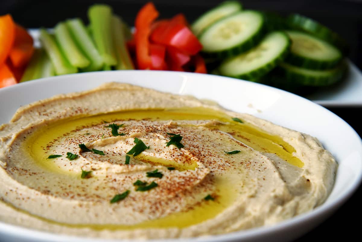 Bowl of hummus drizzled with olive oil and sprinkled with paprika and parsley, with fresh vegetable sticks in the background.