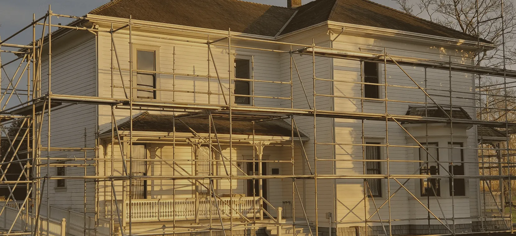 Two-story white house surrounded by metal scaffolding for renovation, illuminated by warm sunlight.