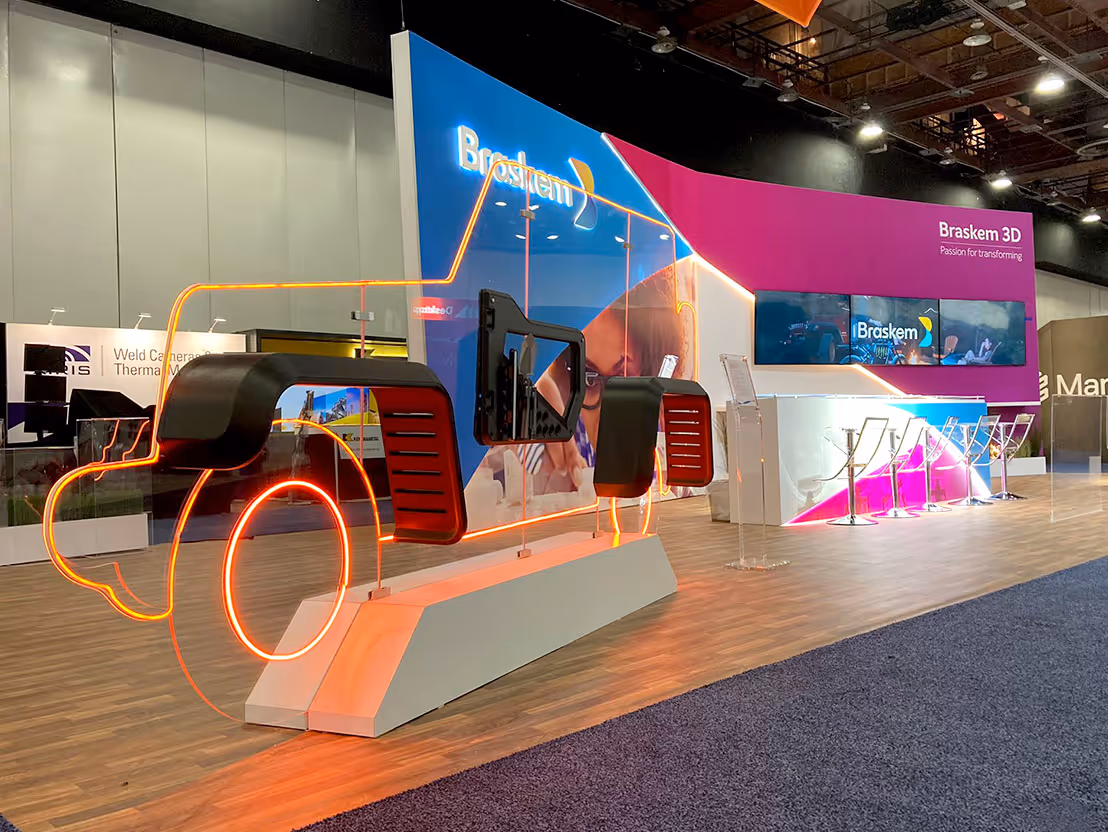 Exhibition booth featuring a neon-outlined truck frame with attached black and red vehicle parts and a backdrop displaying Braskem 3D branding.