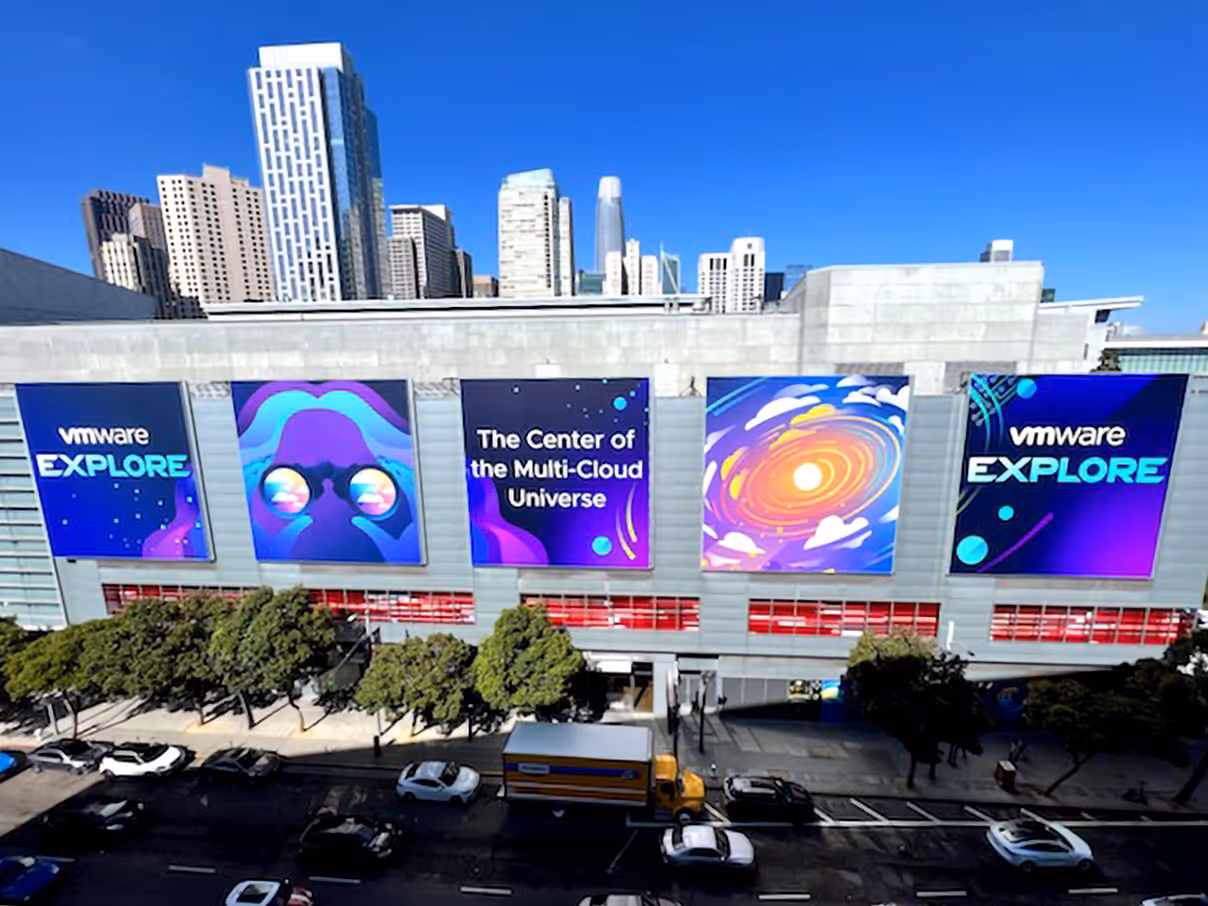 Facade of a convention center with banners displaying 'vmware EXPLORE', an abstract galaxy illustration, and text reading 'The Center of the Multi-Cloud Universe', with a city skyline in the background under clear blue sky.