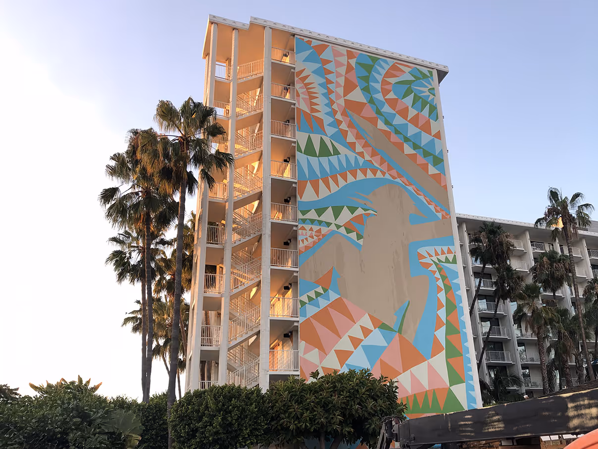 Tall building with an outdoor staircase lit by sunset and a colorful geometric mural on one side, surrounded by palm trees and bushes.