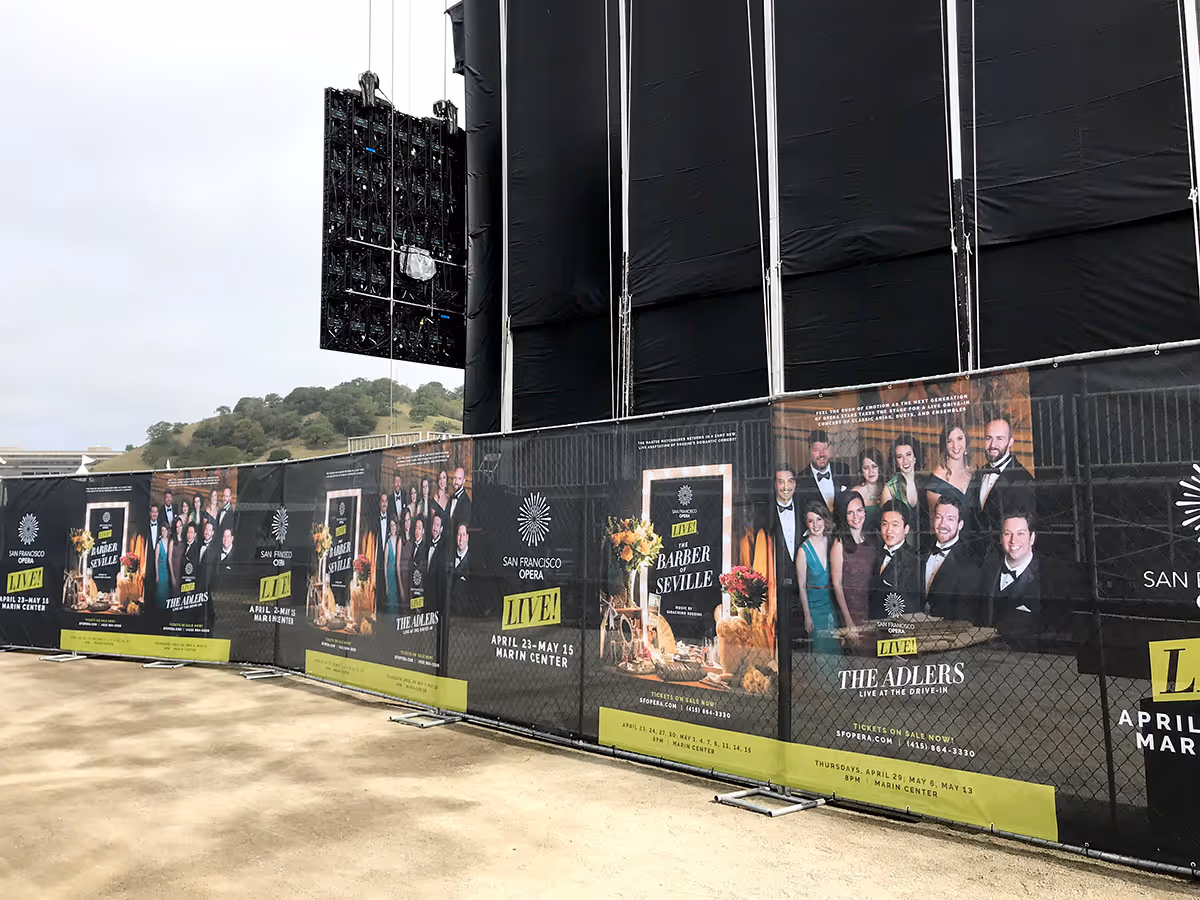 A fenced outdoor stage area featuring large black speaker panels and banners advertising San Francisco Opera LIVE! performances including The Barber of Seville and The Adlers at Marin Center.