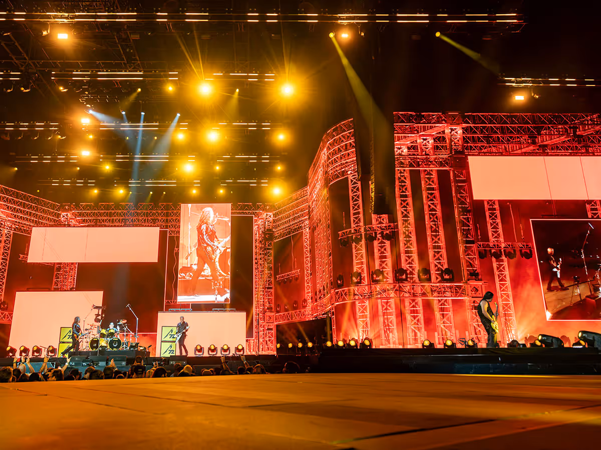 Wide view of a rock band performing on a large, illuminated stage with multiple screens and bright yellow spotlights.