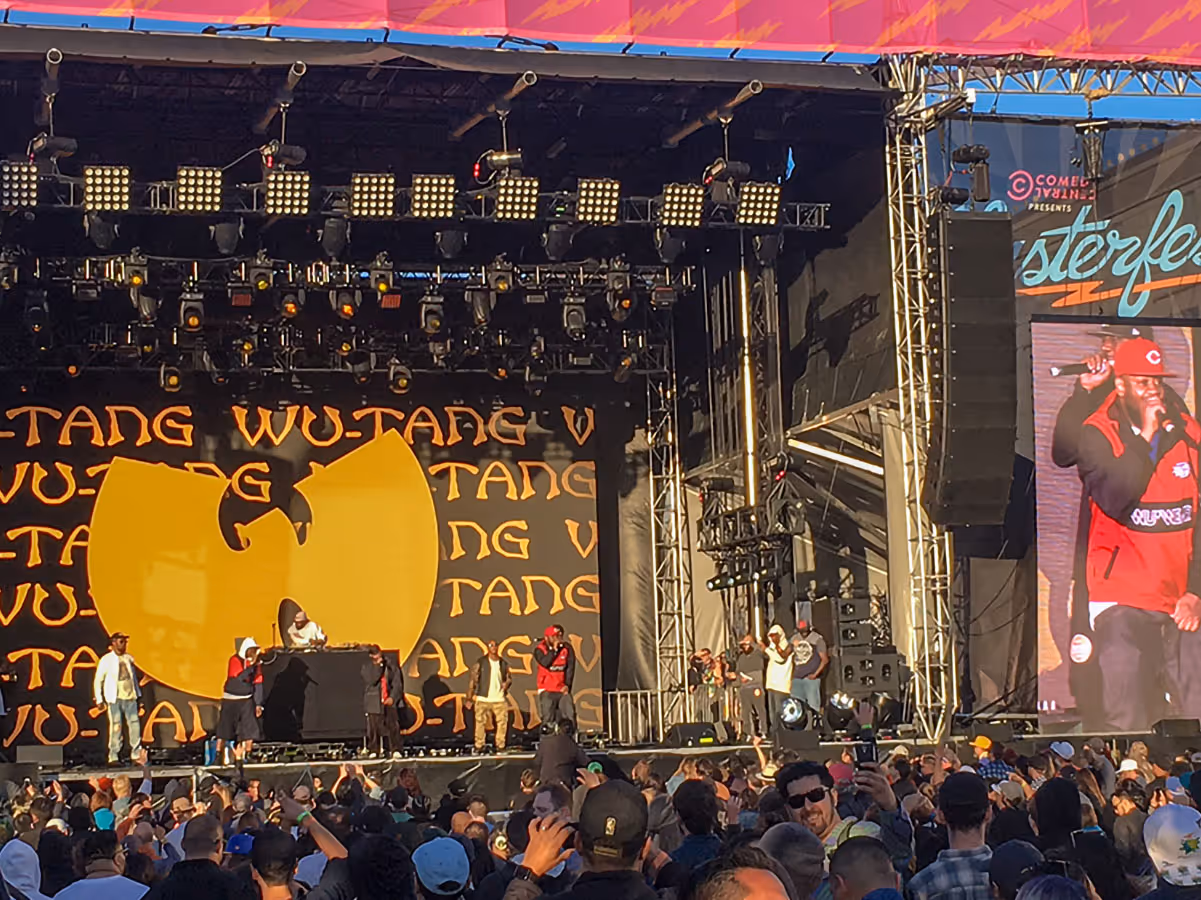 Wu-Tang Clan performing on stage at an outdoor concert with a large crowd watching and the group's logo displayed behind them.