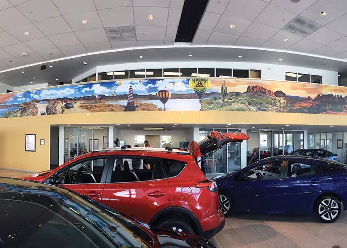 Car dealership showroom interior with red and blue cars parked inside and a panoramic desert-themed mural on the upper wall.