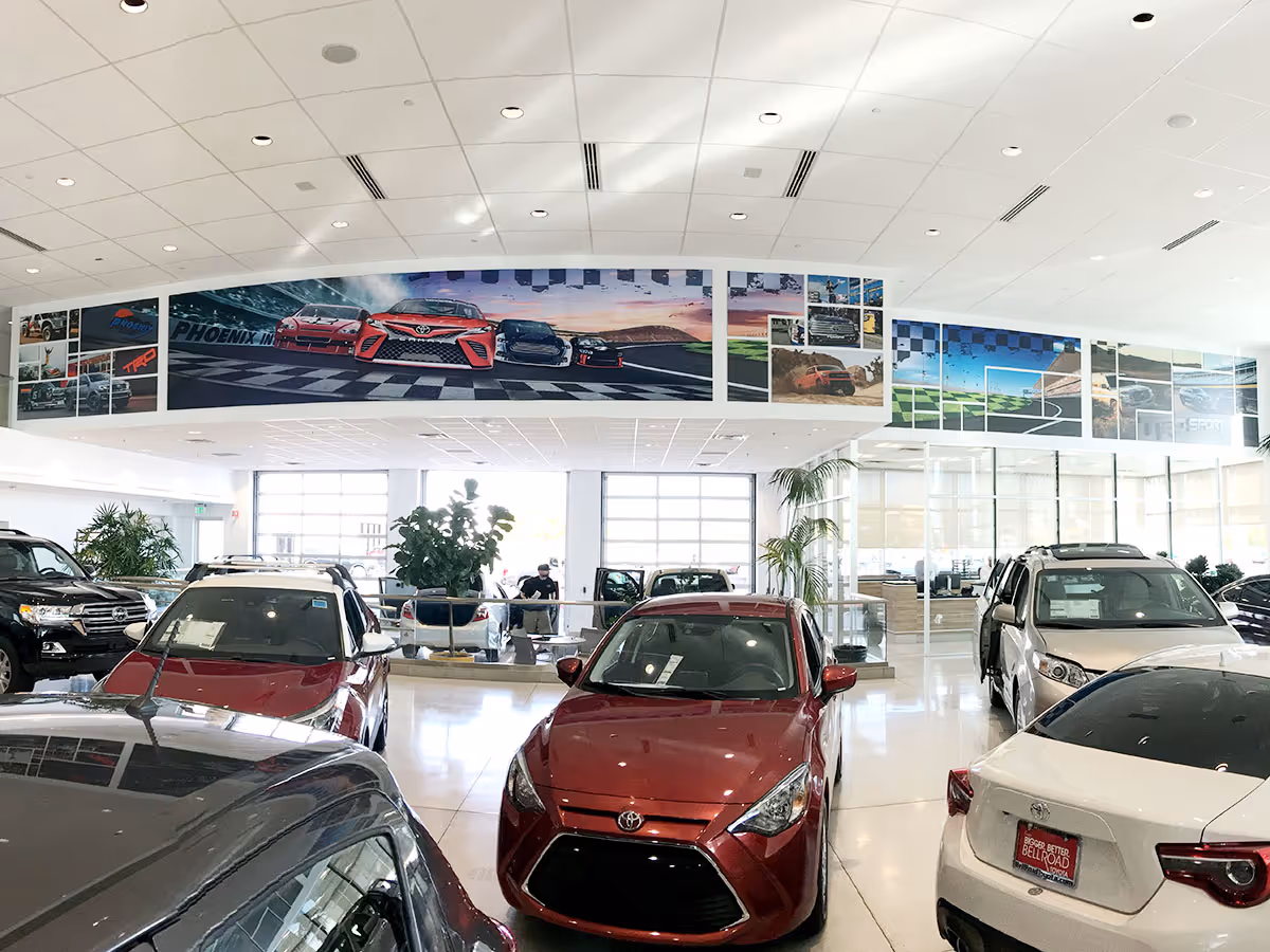 Interior of a bright car dealership showroom featuring various Toyota vehicles and racing-themed wall art above.