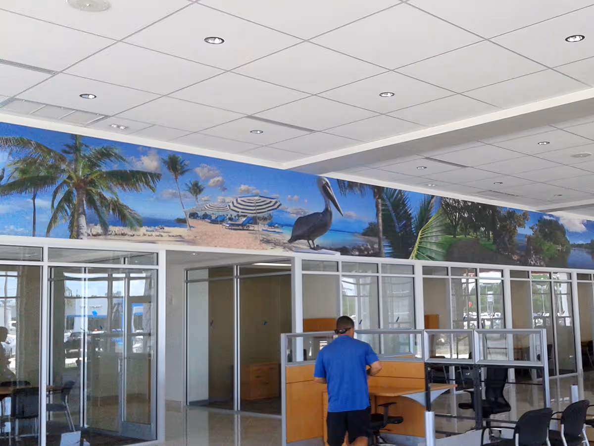 Interior office space with glass-walled rooms, cubicles, and a person in a blue shirt; a tropical beach and pelican mural runs along the top wall.