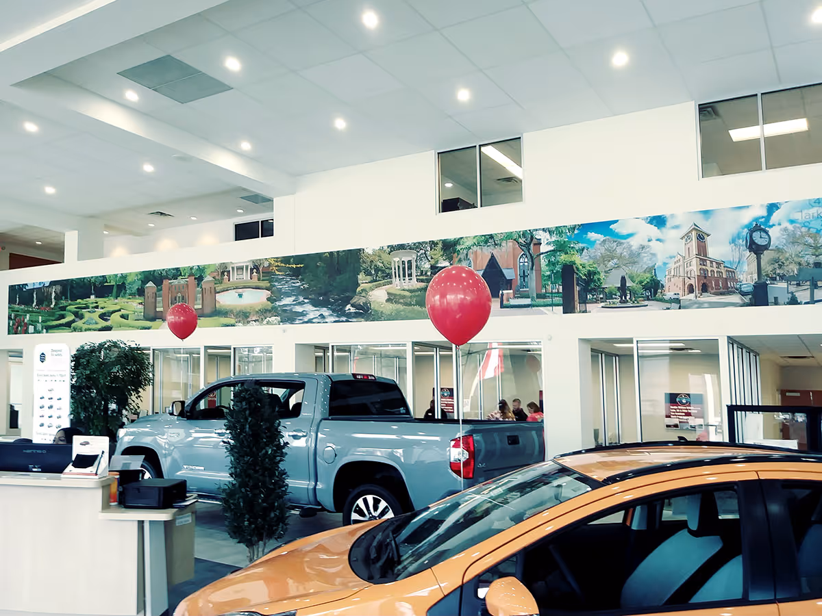 Interior of a showroom with a blue pickup truck and an orange car, decorated with red balloons and a mural of outdoor scenes on the wall.