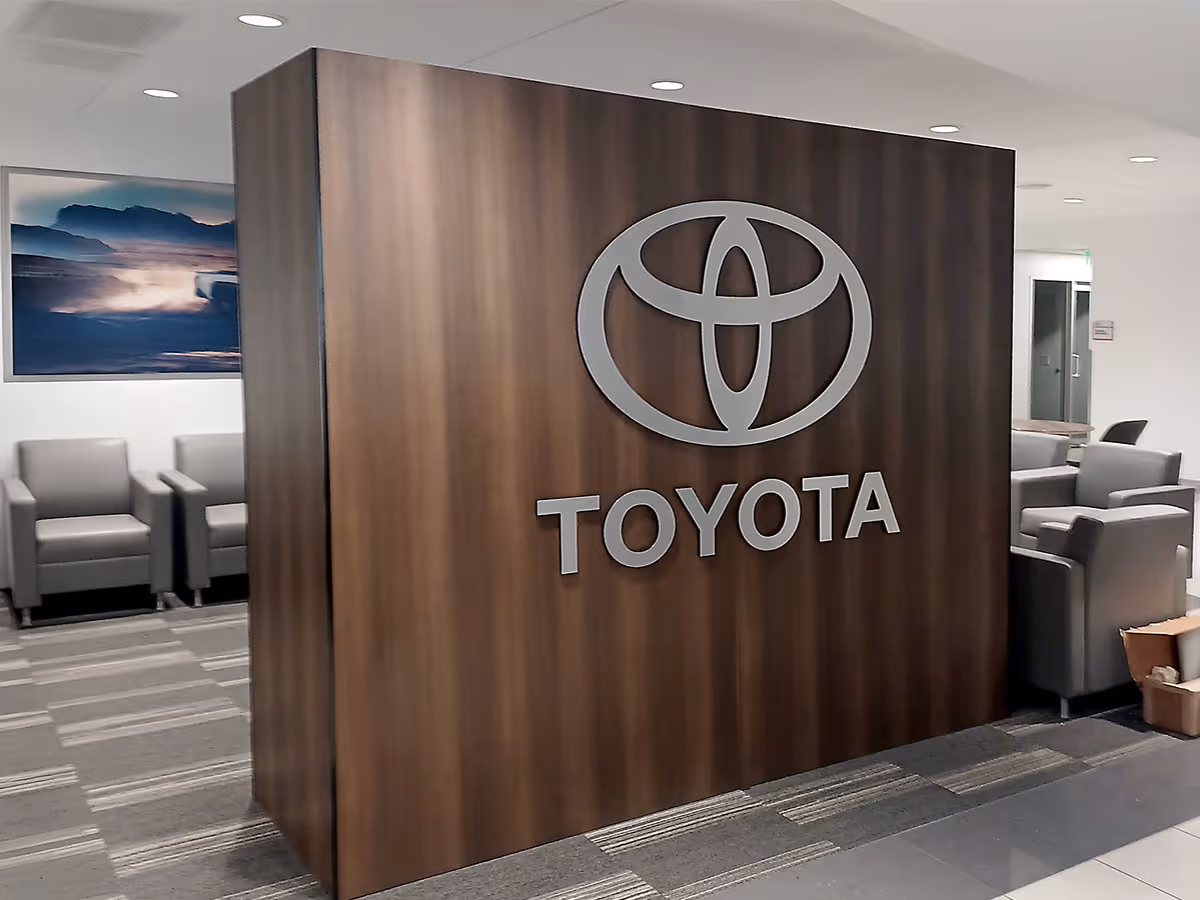 Wood-paneled wall with a large fabricated silver Toyota logo in a modern office lounge area with grey armchairs.