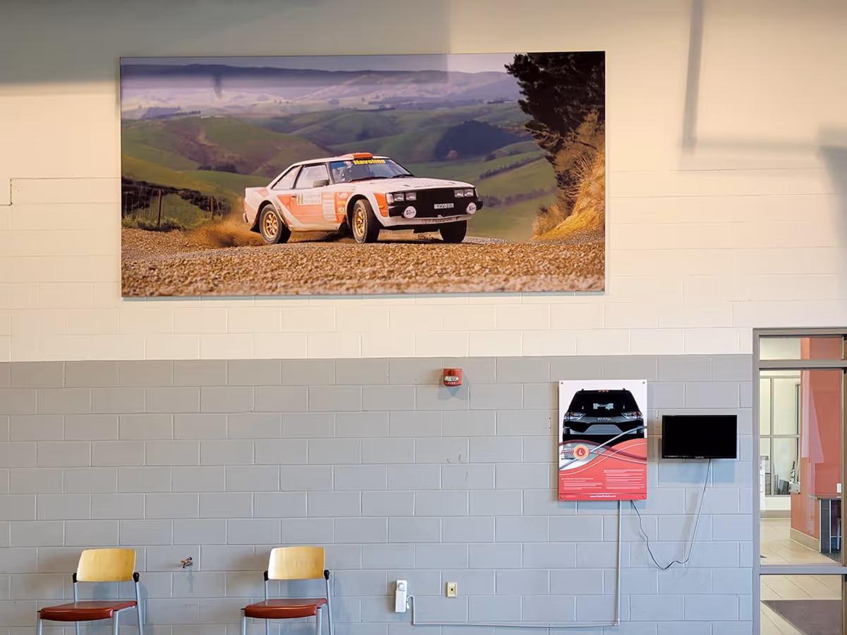 Interior wall of a room with two chairs below a large Sicicone edged Graphic frame with a photo of a rally car driving on a gravel road in hilly countryside.
