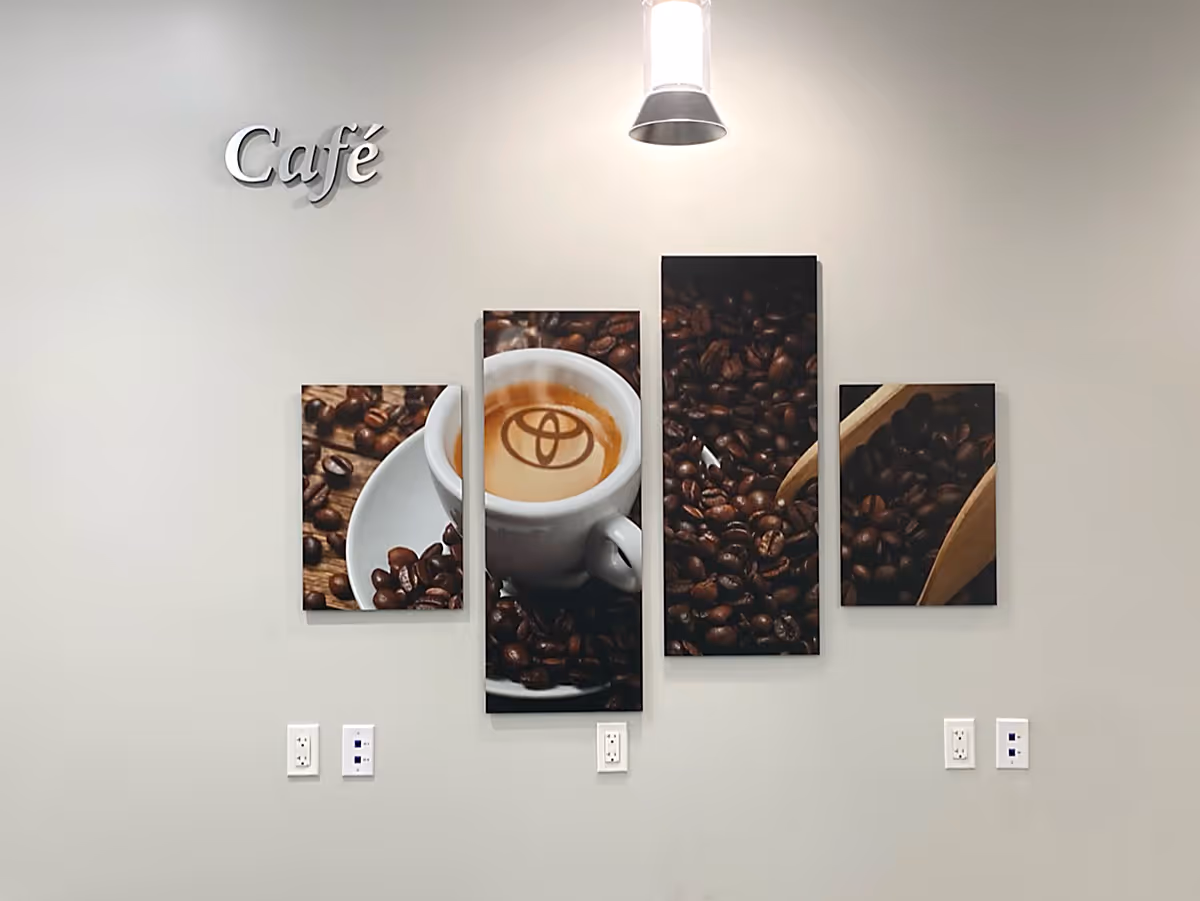 Four-panel Silicone Edged Graphic Frames with Fabric wall art showing a cup of coffee with a Toyota logo in the foam surrounded by coffee beans and wooden scoops; a silver café sign is mounted above.