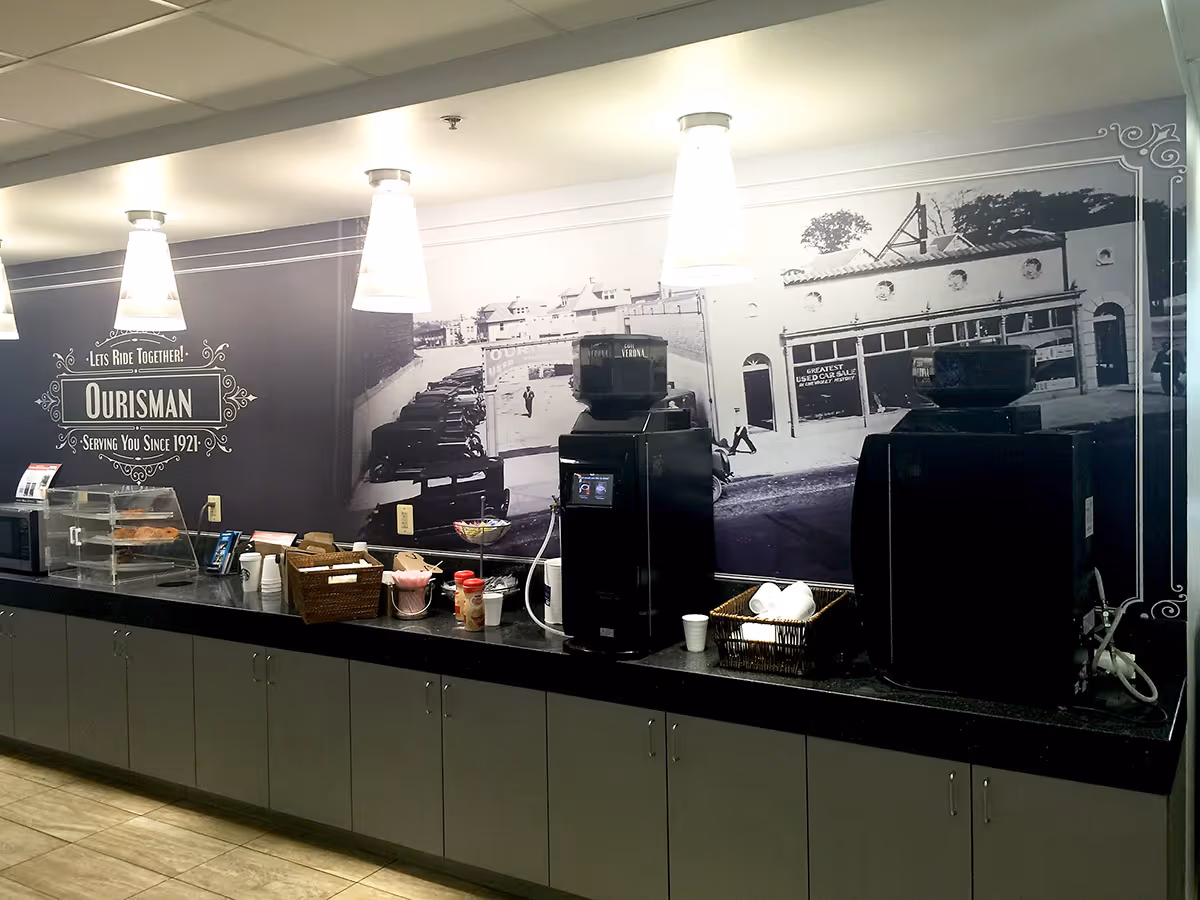 Coffee station with two black coffee dispensers, cups, condiments, and pastries in a modern kitchen area with a black-and-white vintage wall mural decal and hanging pendant lights.