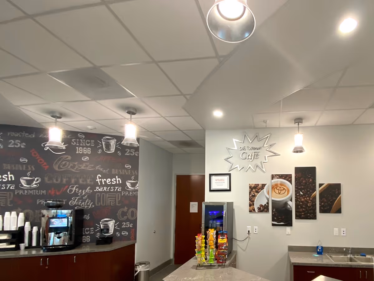 Modern office café area with coffee machine, snack display, and wall decal art featuring coffee beans and a cup with a Toyota logo in the foam. Wall on the right showcases sign and Fabric art.