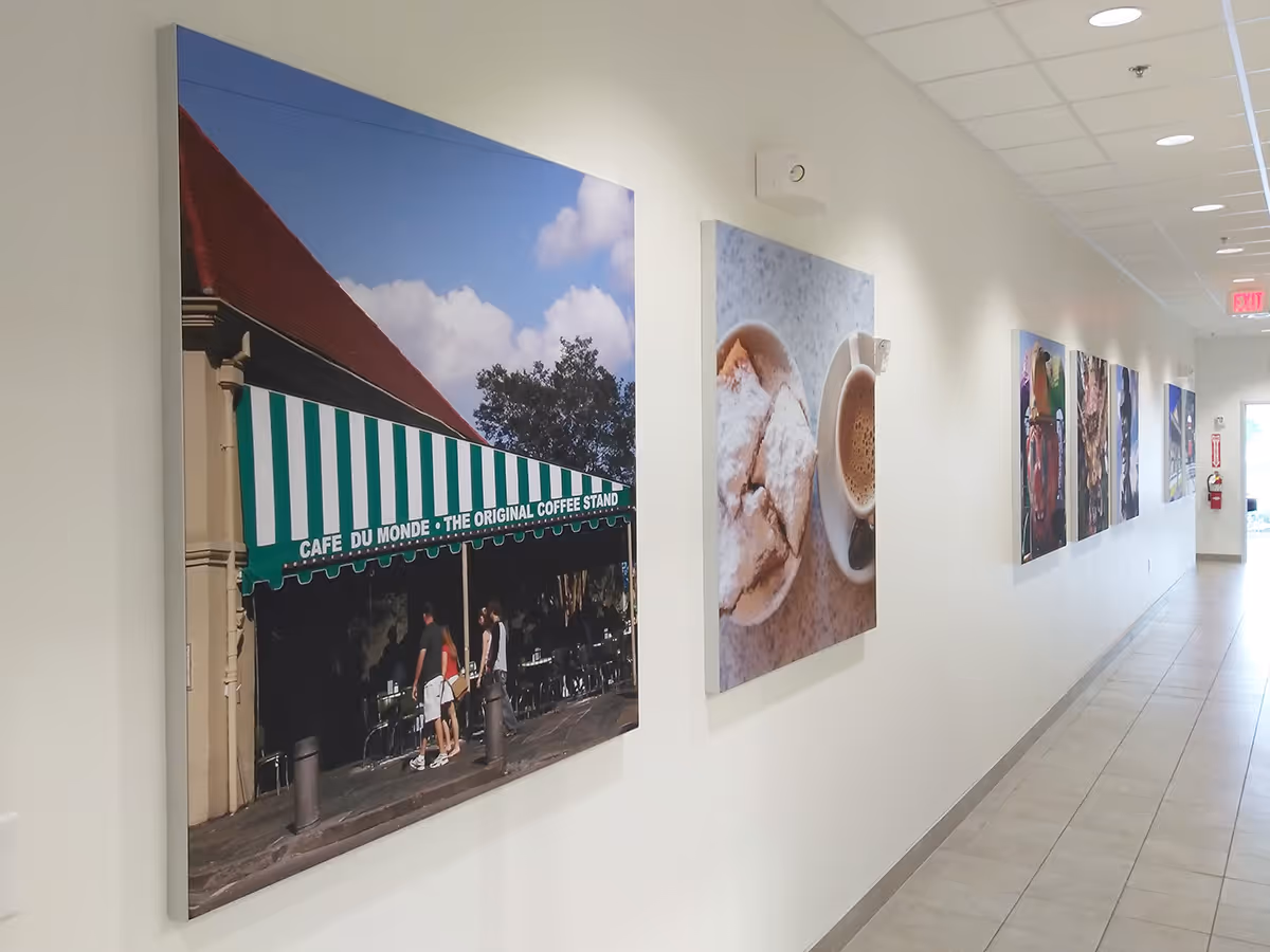 A hallway with a series of large silicone edged graphic framed fabric prints on one side, including one of a Café du Monde coffee stand with people walking by and another of a cup of coffee and pastries.