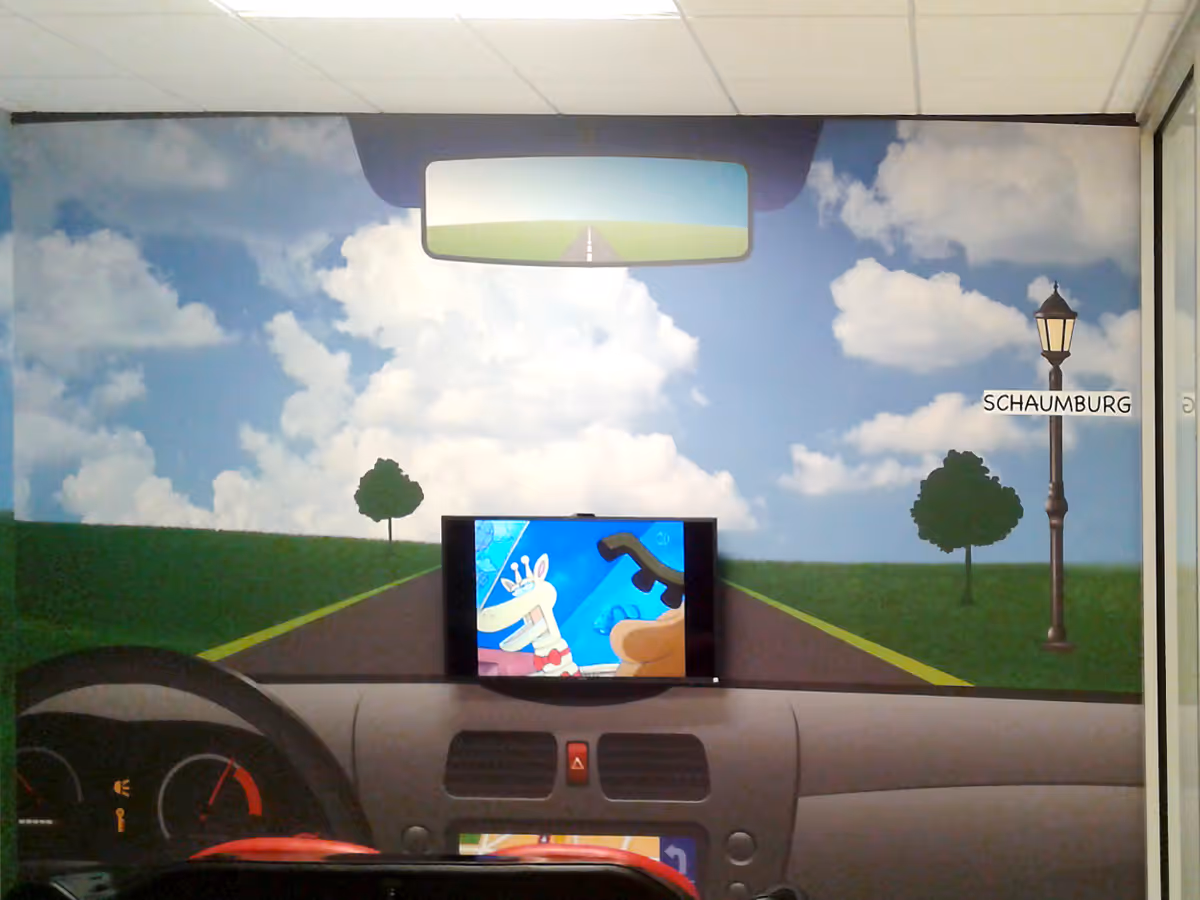 Wall mural showing a car dashboard and steering wheel with a road extending ahead under a blue sky with clouds, featuring a rearview mirror and a street lamp with a Schaumburg sign, and a small screen displaying a cartoon giraffe inside the car.